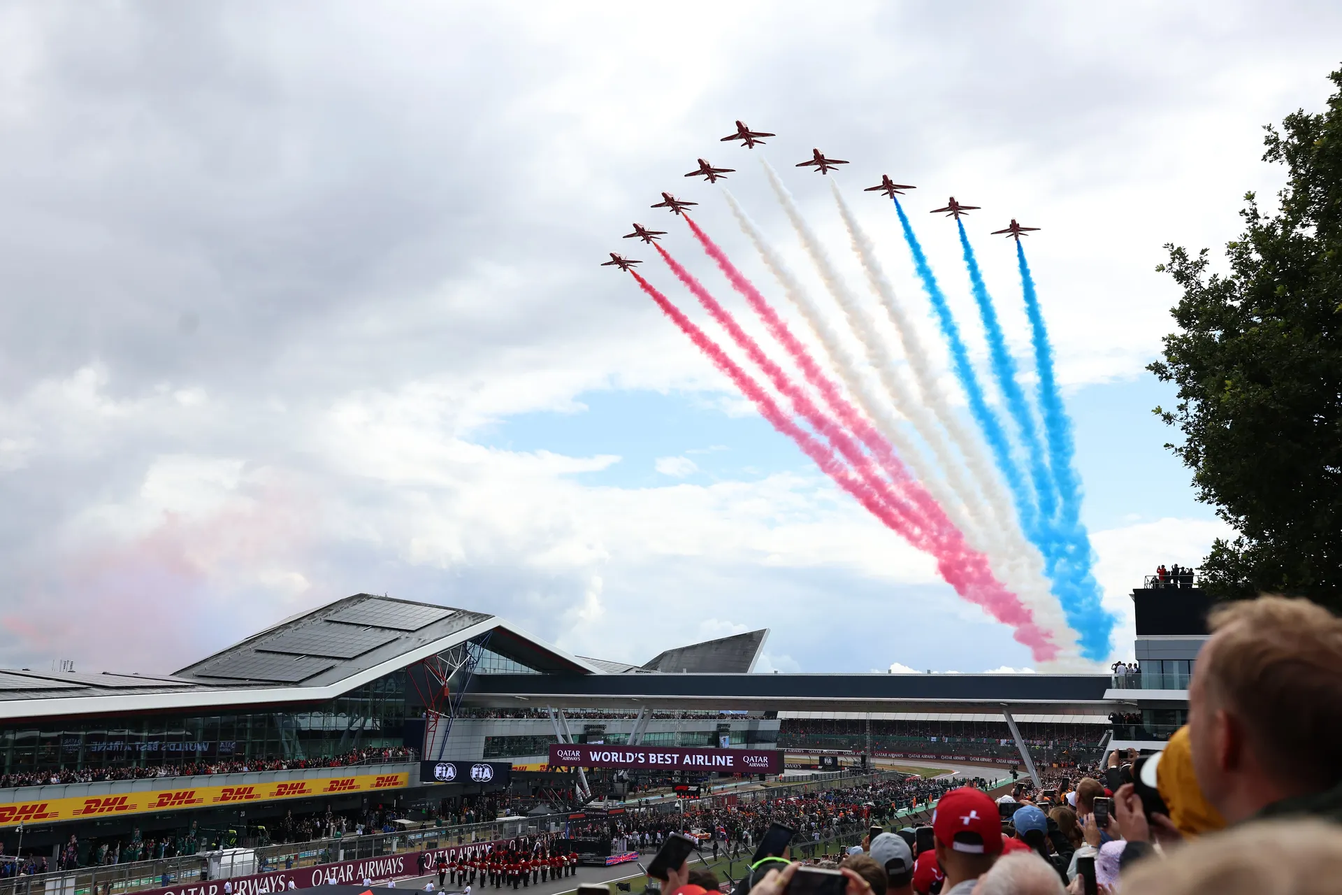 Aerial display jets sweep across the sky above Silverstone during the British Grand Prix, trailing red, white and blue smoke over the packed main straight and grandstands, marking the start of race day atmosphere.