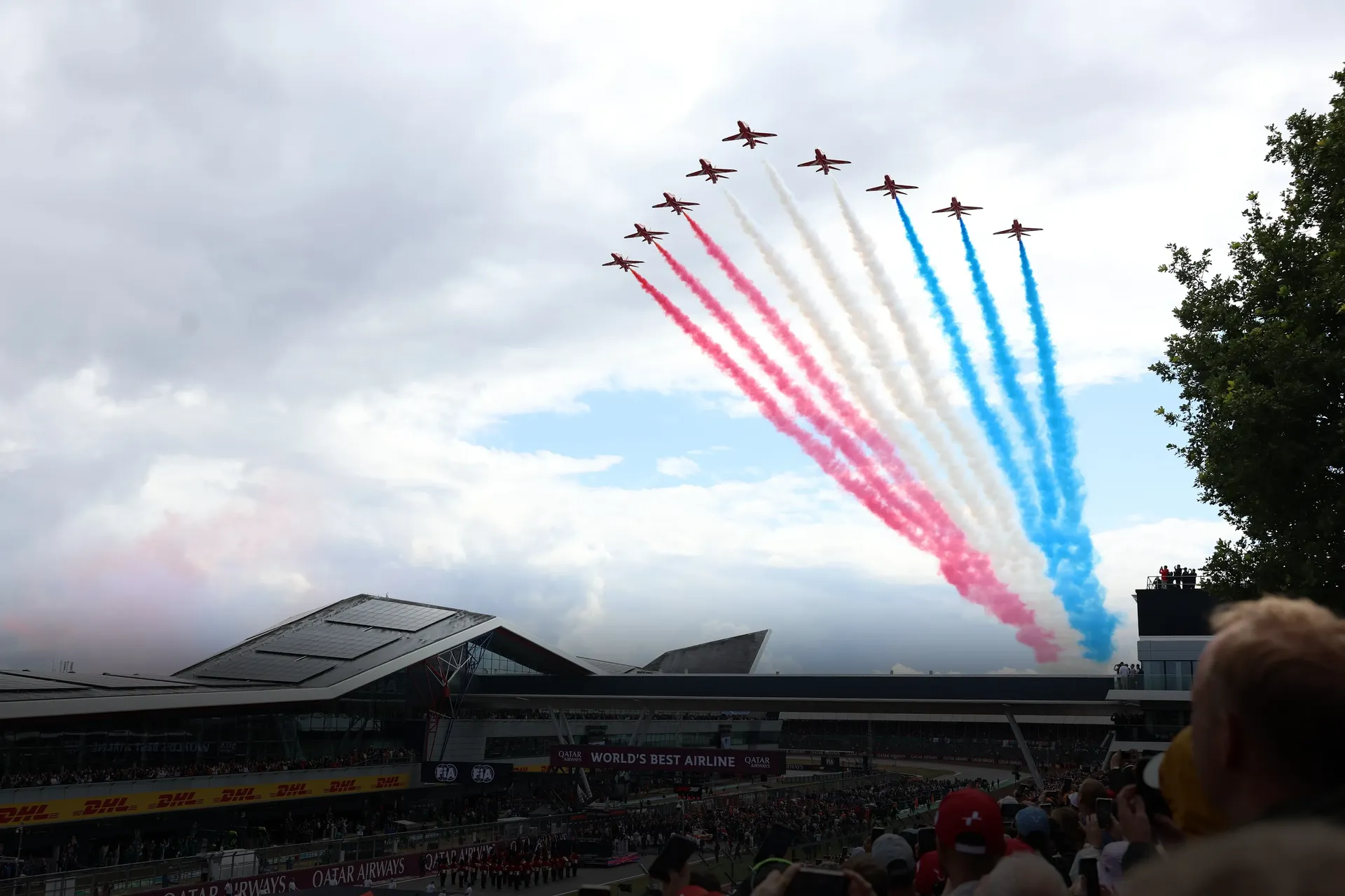 Aerial display jets sweep across the sky above Silverstone during the British Grand Prix, trailing red, white and blue smoke over the packed main straight and grandstands, marking the start of race day atmosphere.