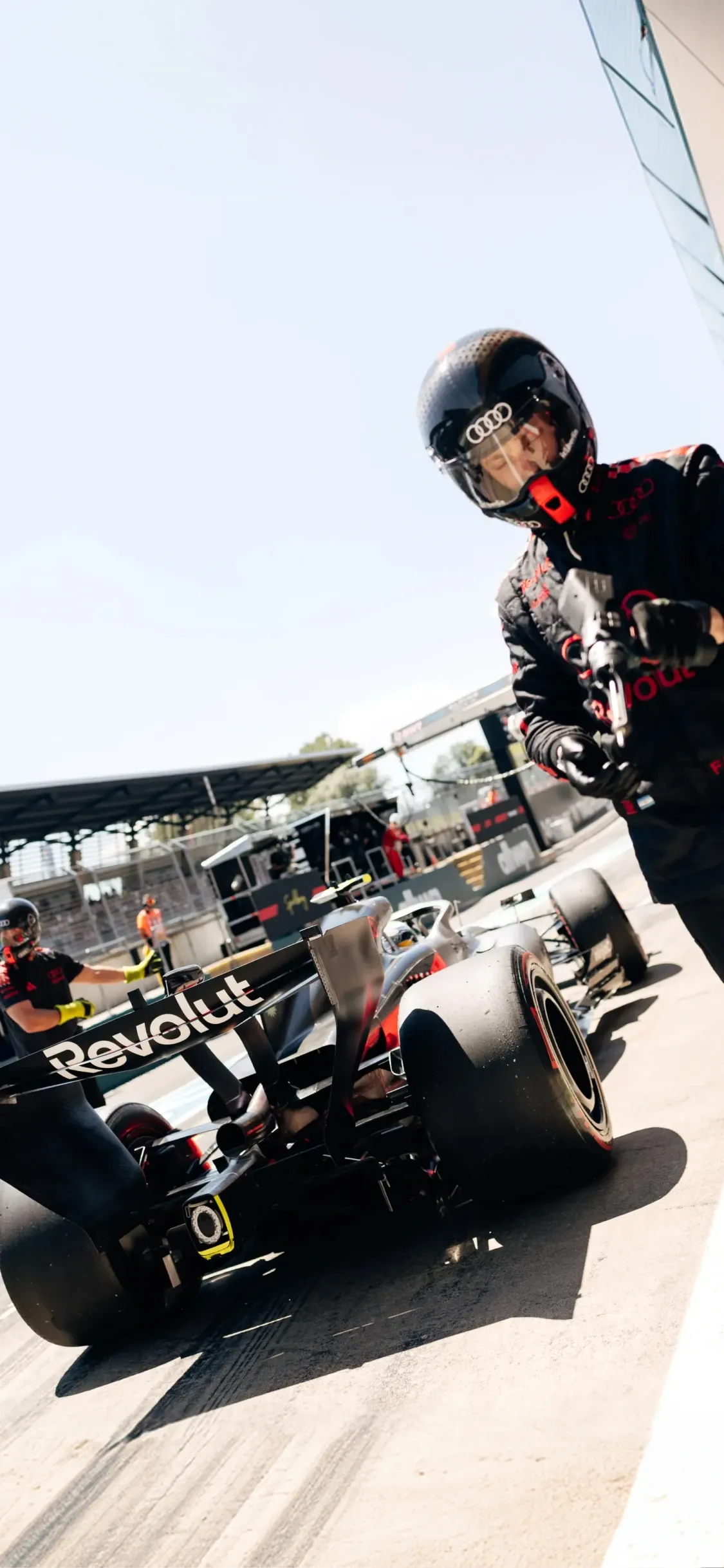Audi Revolut F1® Team pit crew prepares the car in the pit lane, one mechanic stepping forward with equipment as the car is readied under bright daylight.
