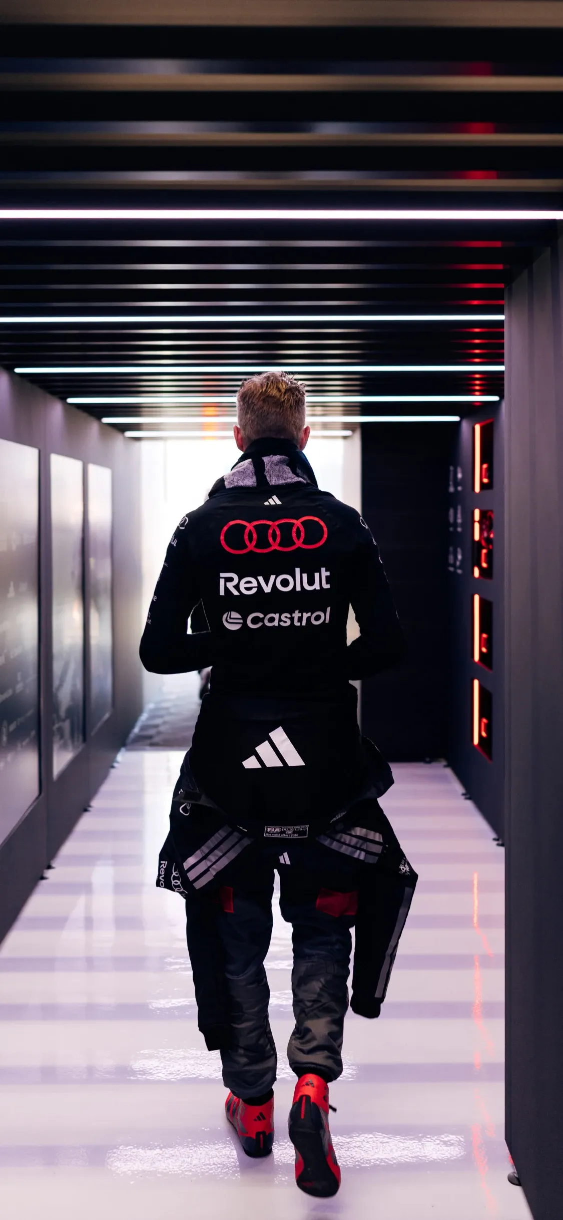 Audi Revolut F1® Team driver Nico Hulkenberg walking down a lit paddock corridor from behind, heading towards the garage in full race gear.