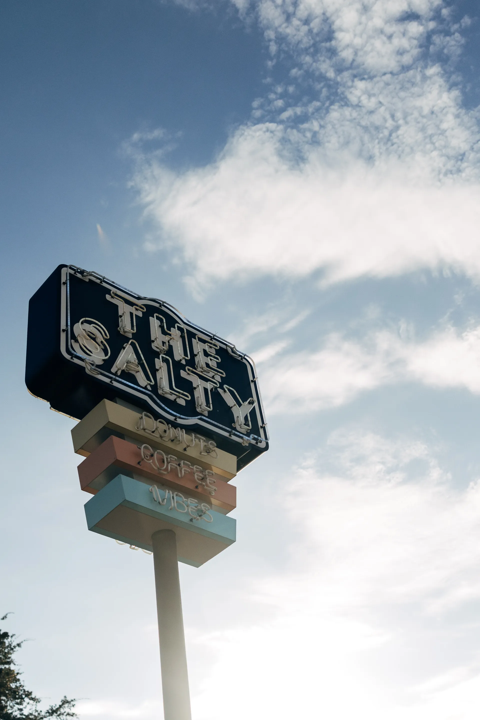 Neon sign of The Salty reading Donuts, Coffee, Vibes against a cloudy sky in Austin
