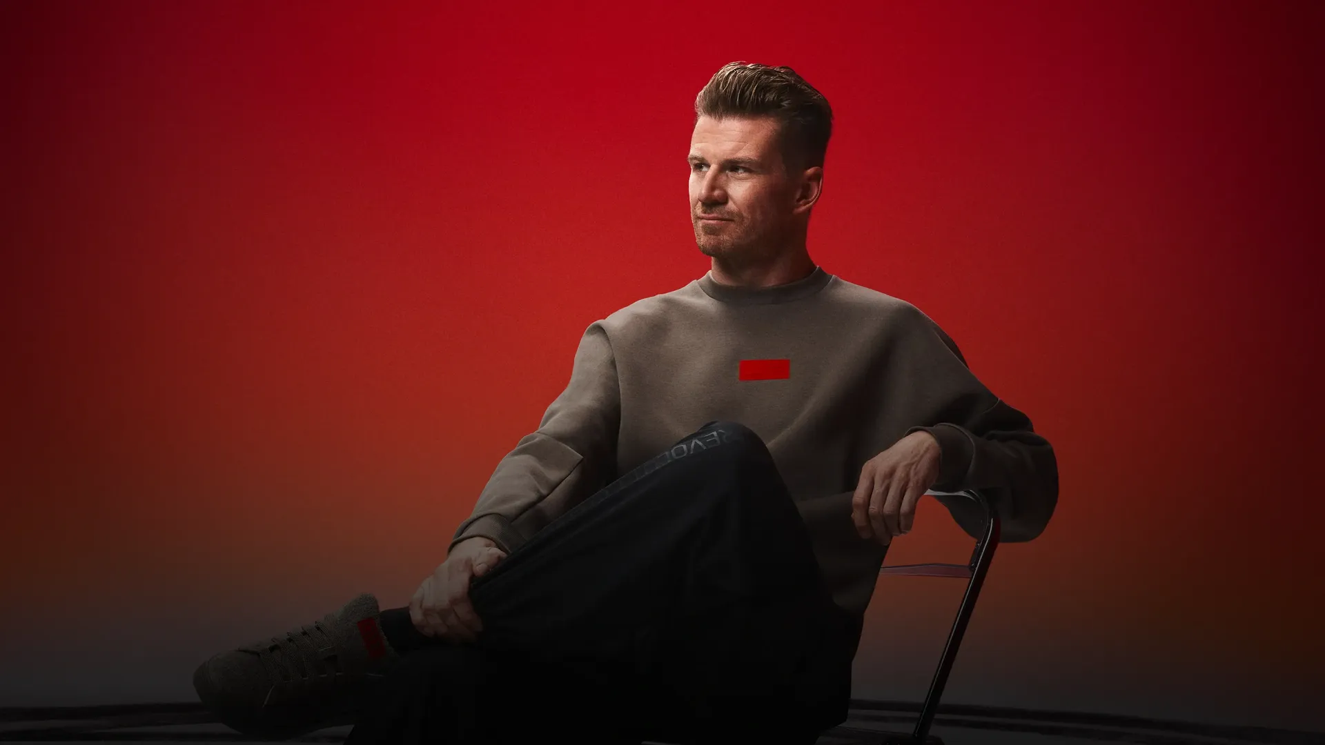 Nico Hulkenberg wearing adidas x Audi Revolut F1® Team Fanwear collection graphic sweatshirt and woven pants, sittin on a stool in the studio