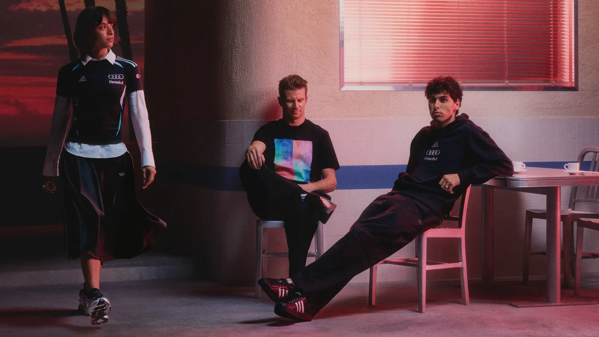 Young female model walking past on the left, while Nico Hülkenberg and Gabriel Bortoleto sit side by side on stools on the right in a relaxed pose, wearing casual adidas fanwear in a warmly lit studio setting