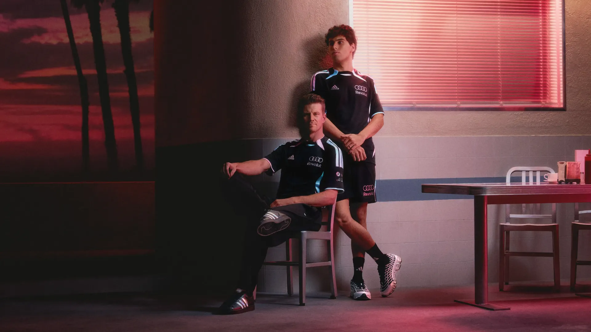 Portrait of Audi Revolut F1® Team drivers Nico Hulkenberg seated on a stool, with Gabriel Bortoleto standing behind him, both in a relaxed pose wearing black teamwear under warm studio lighting