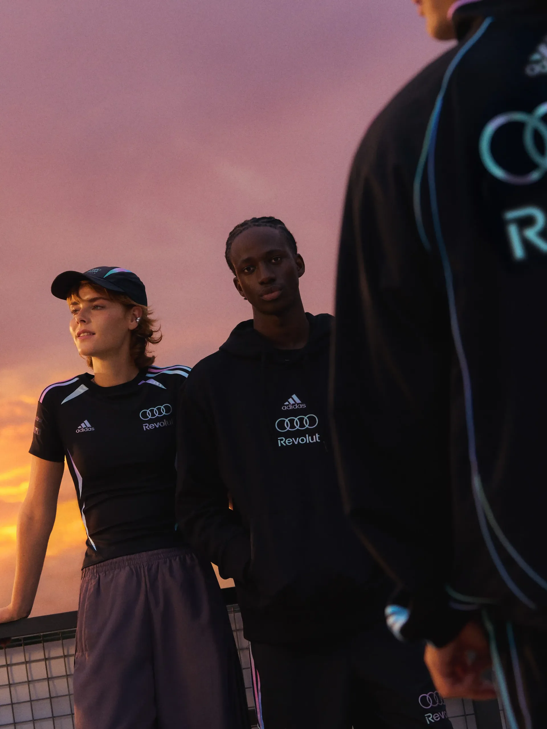 Group of three male and female models standing together in a studio with a sunset-style background, wearing black adidas Miami fanwear outfits