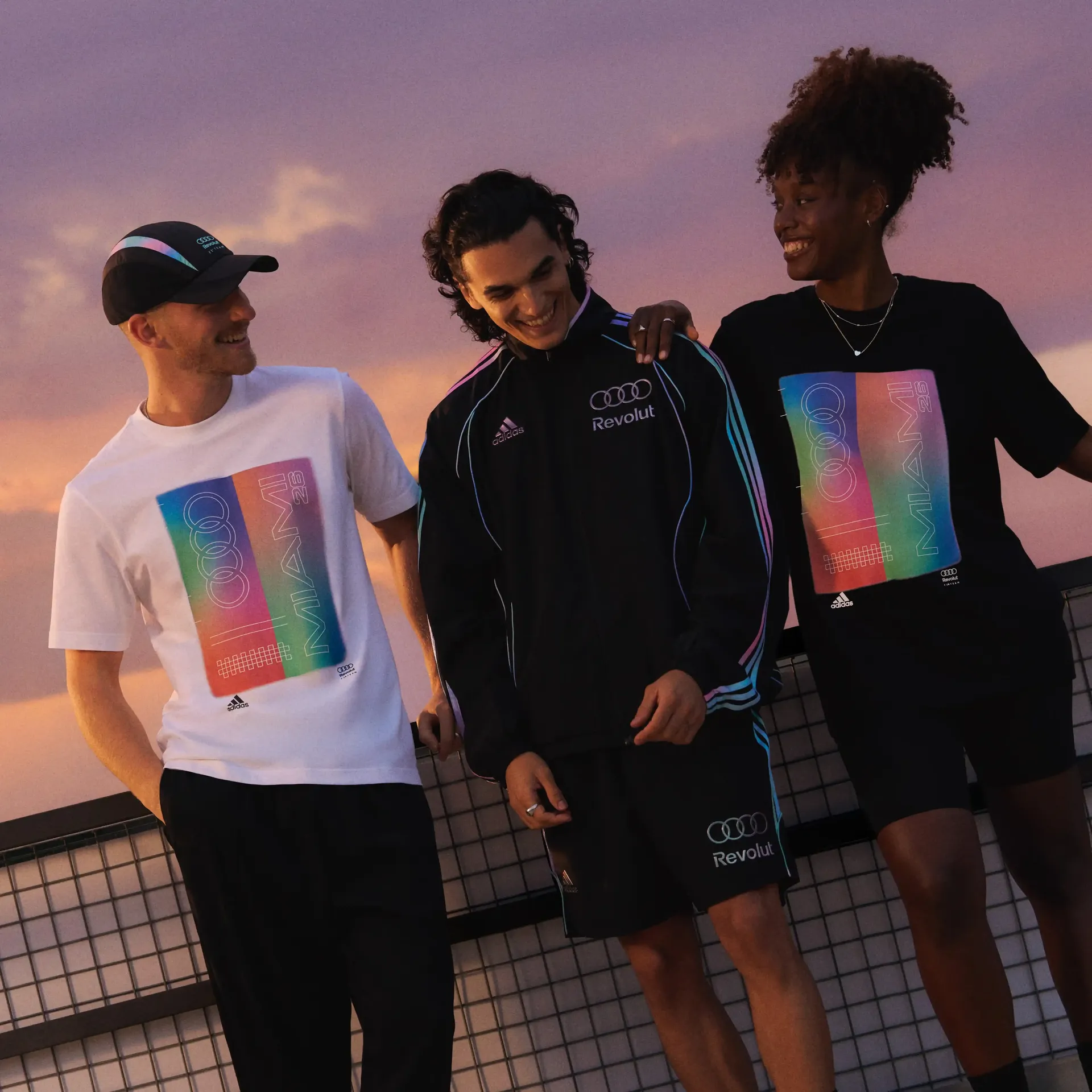 Relaxed group of three young male and female models standing together in a studio with a sunset-style background, wearing casual adidas Miami fanwear outfits