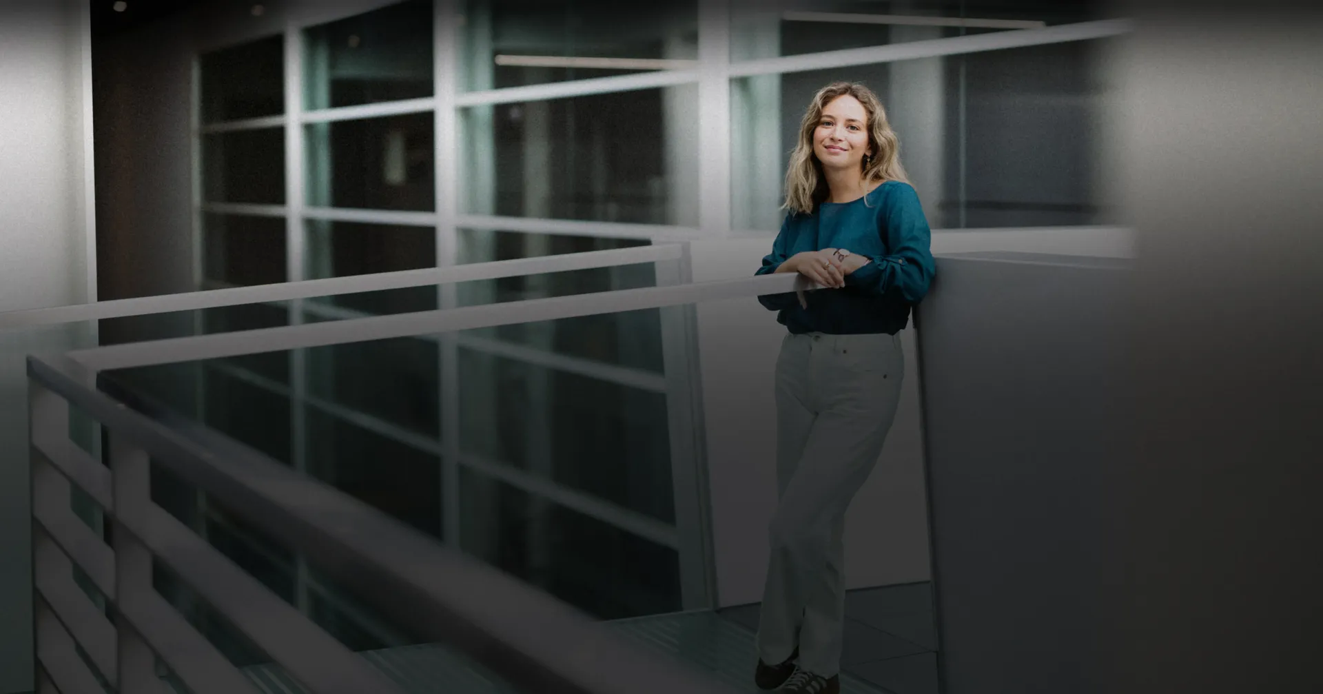 Sofia in a teal top and light trousers, leaning against a railing in a bright, modern office atrium with glass walls and white architectural details.