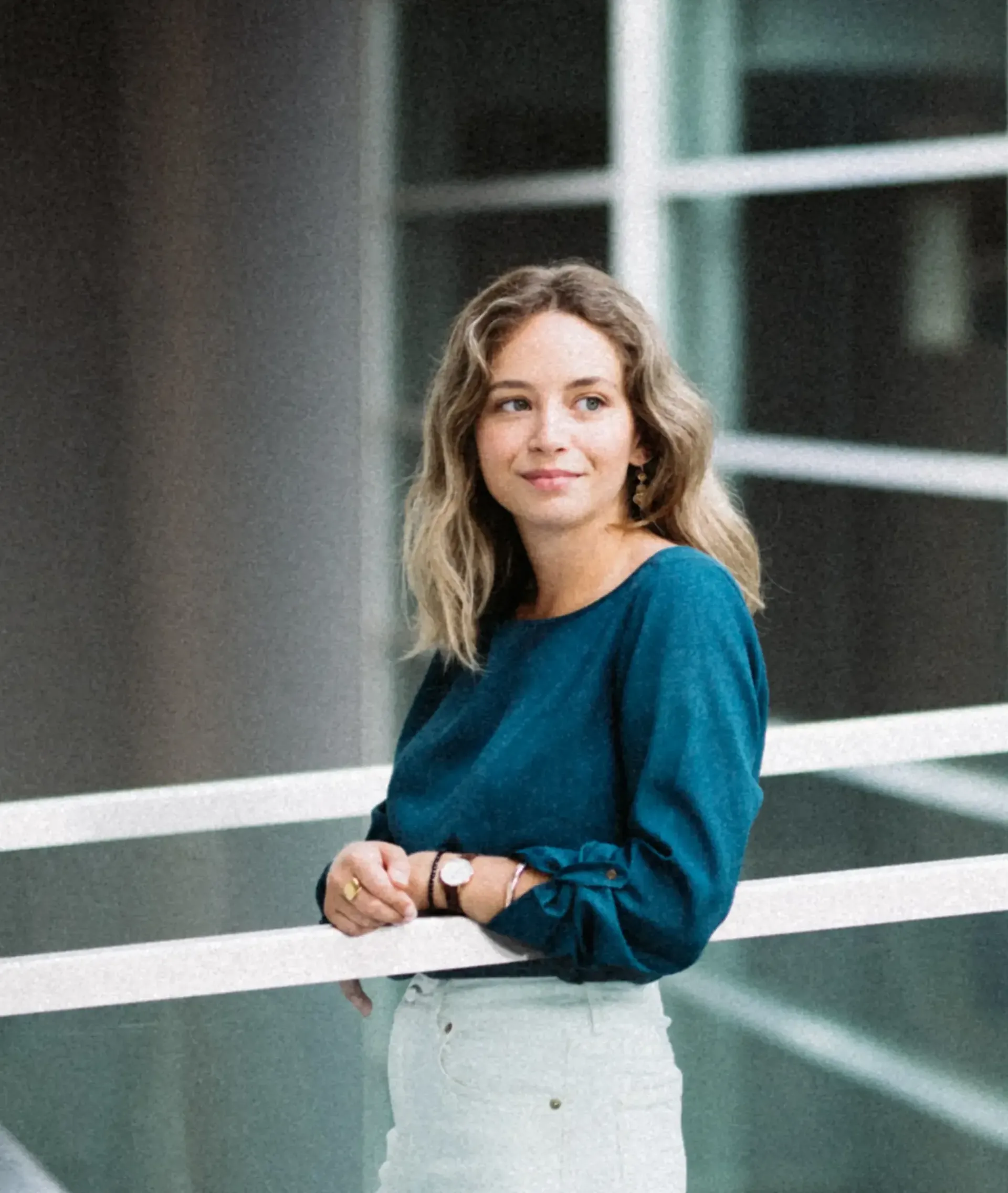 Portrait-format shot of Sofia in a teal top, standing at a railing in a modern office corridor, partly framed by a white column in the foreground.