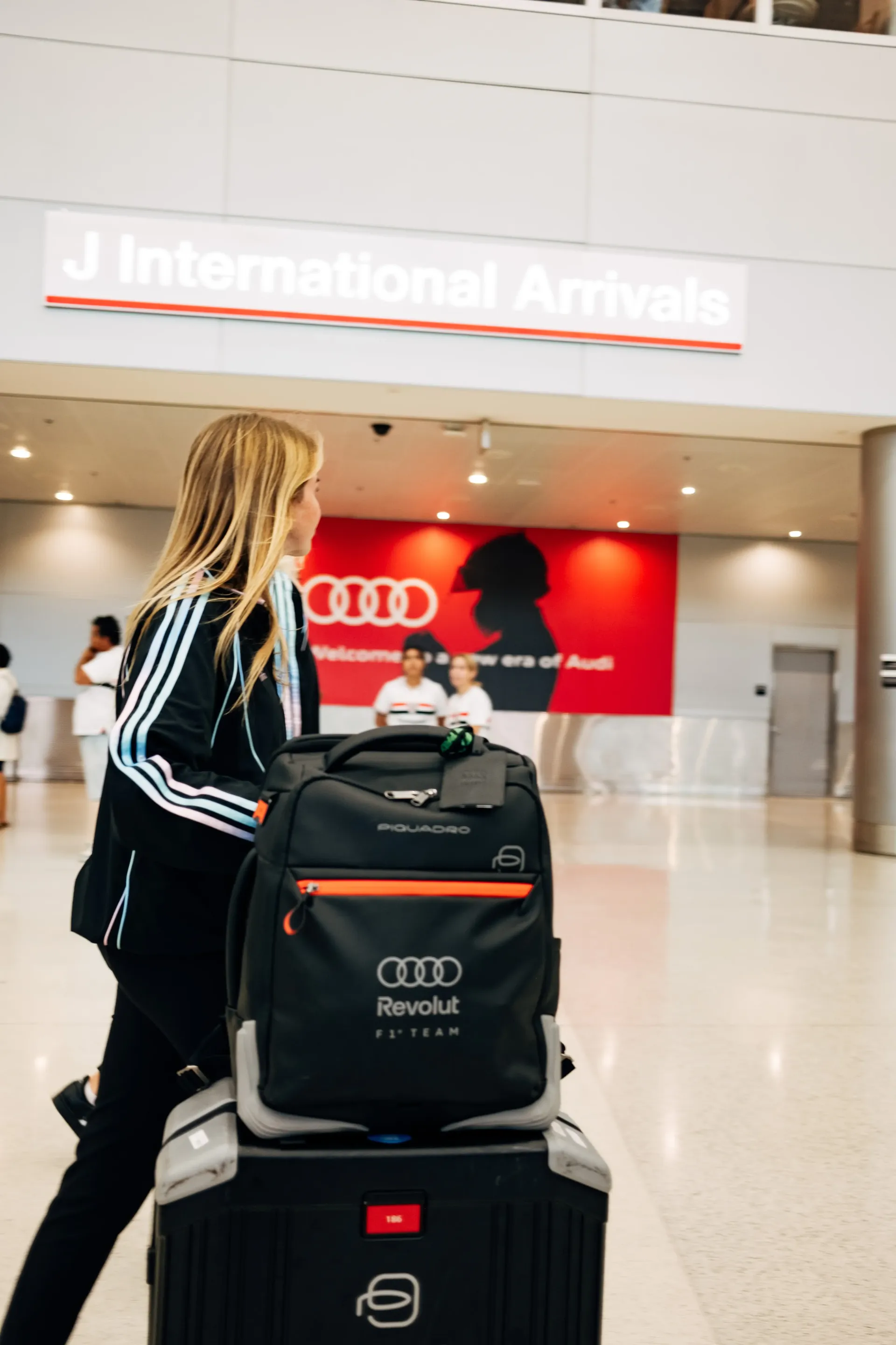 Audi Revolut F1®Team female crew member walking through terminal corridor in the new Miami collection tracksuit and backpacks