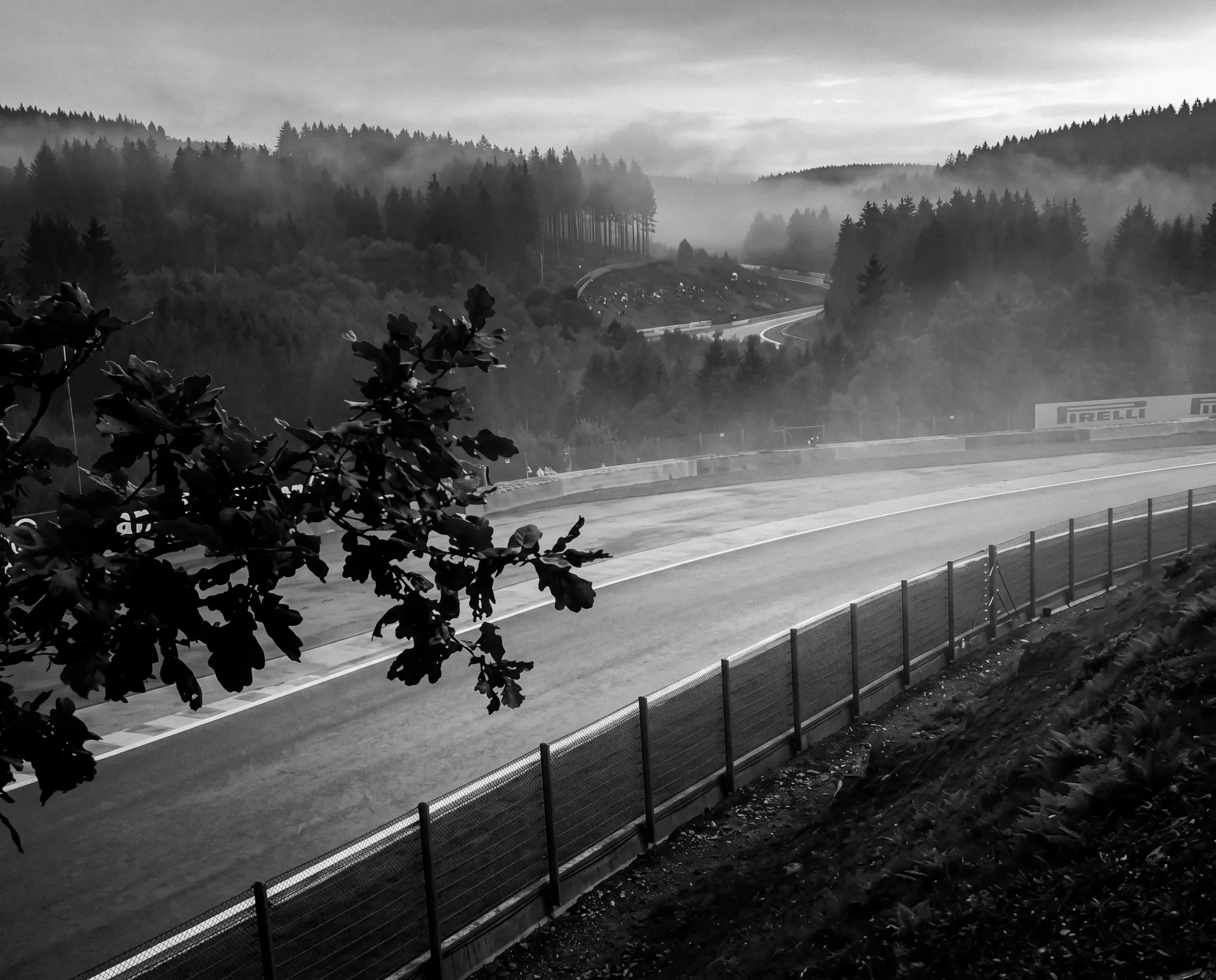 Black-and-white view of the Blanchimont corner at Circuit de Spa-Francorchamps with misty forested hills in the background.