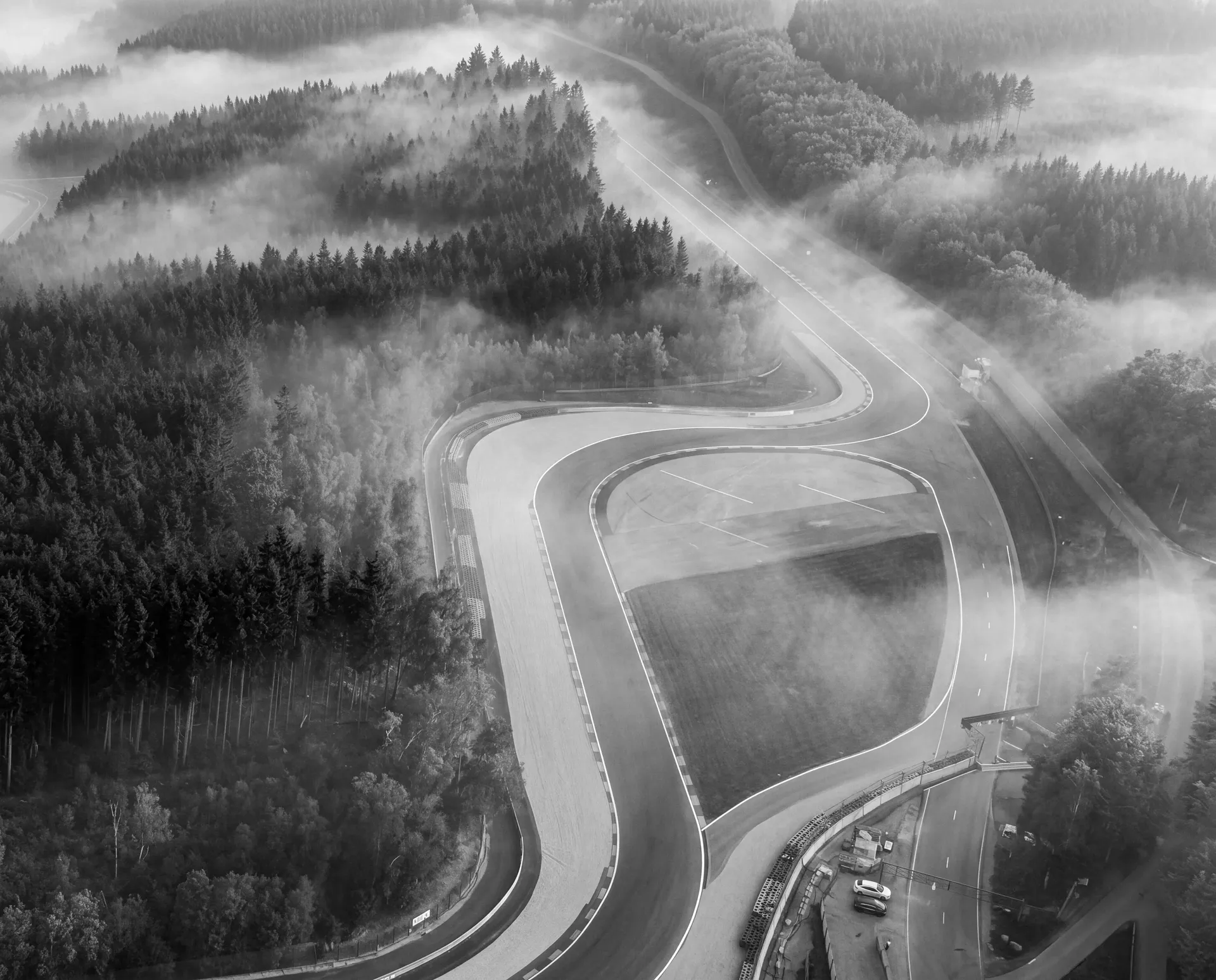 Black-and-white aerial view of the Les Combes section winding through mist-covered forests at Circuit de Spa-Francorchamps.