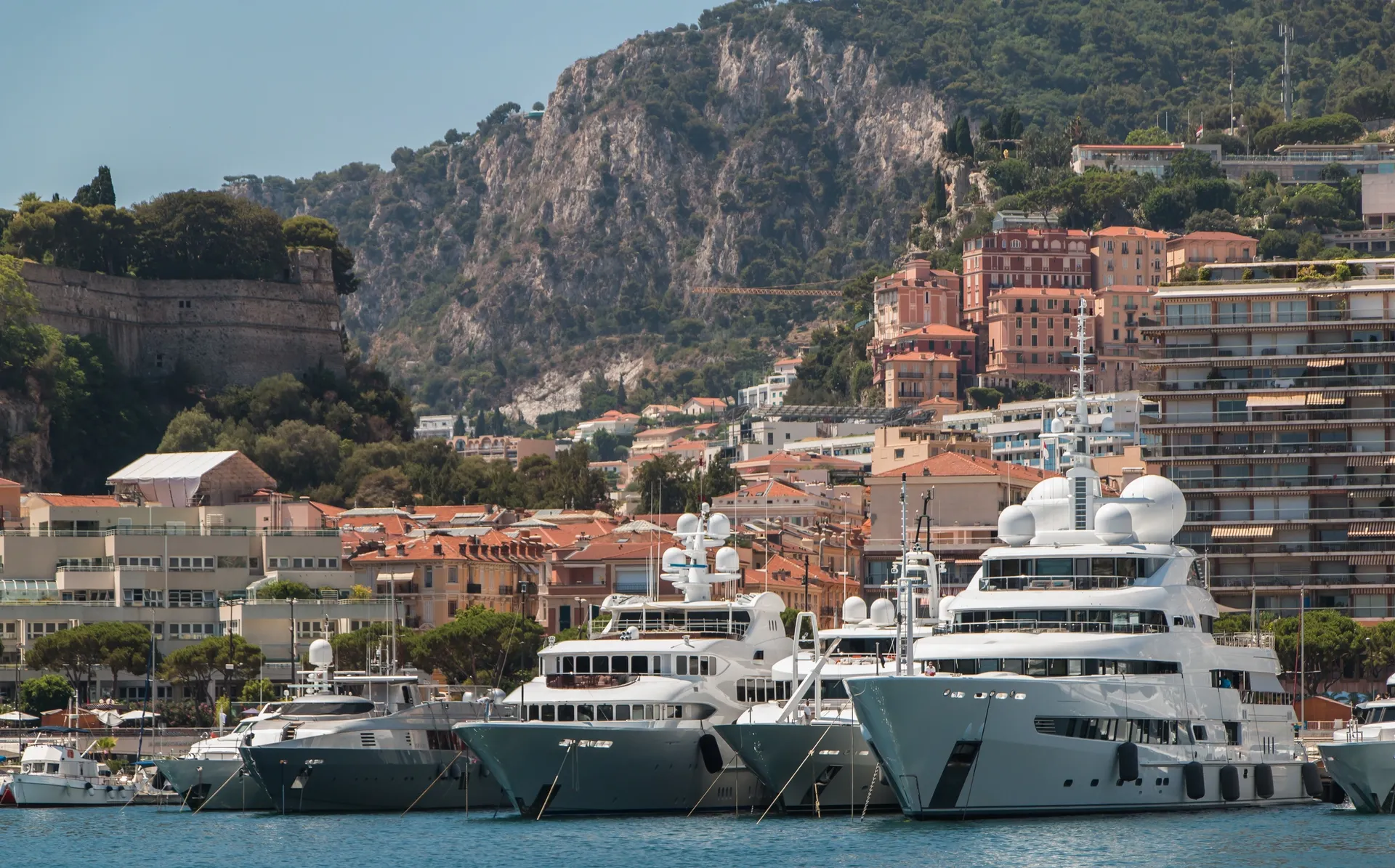Luxury yachts in Monaco's harbor with waterfront buildings and rocky hillsides in the background.