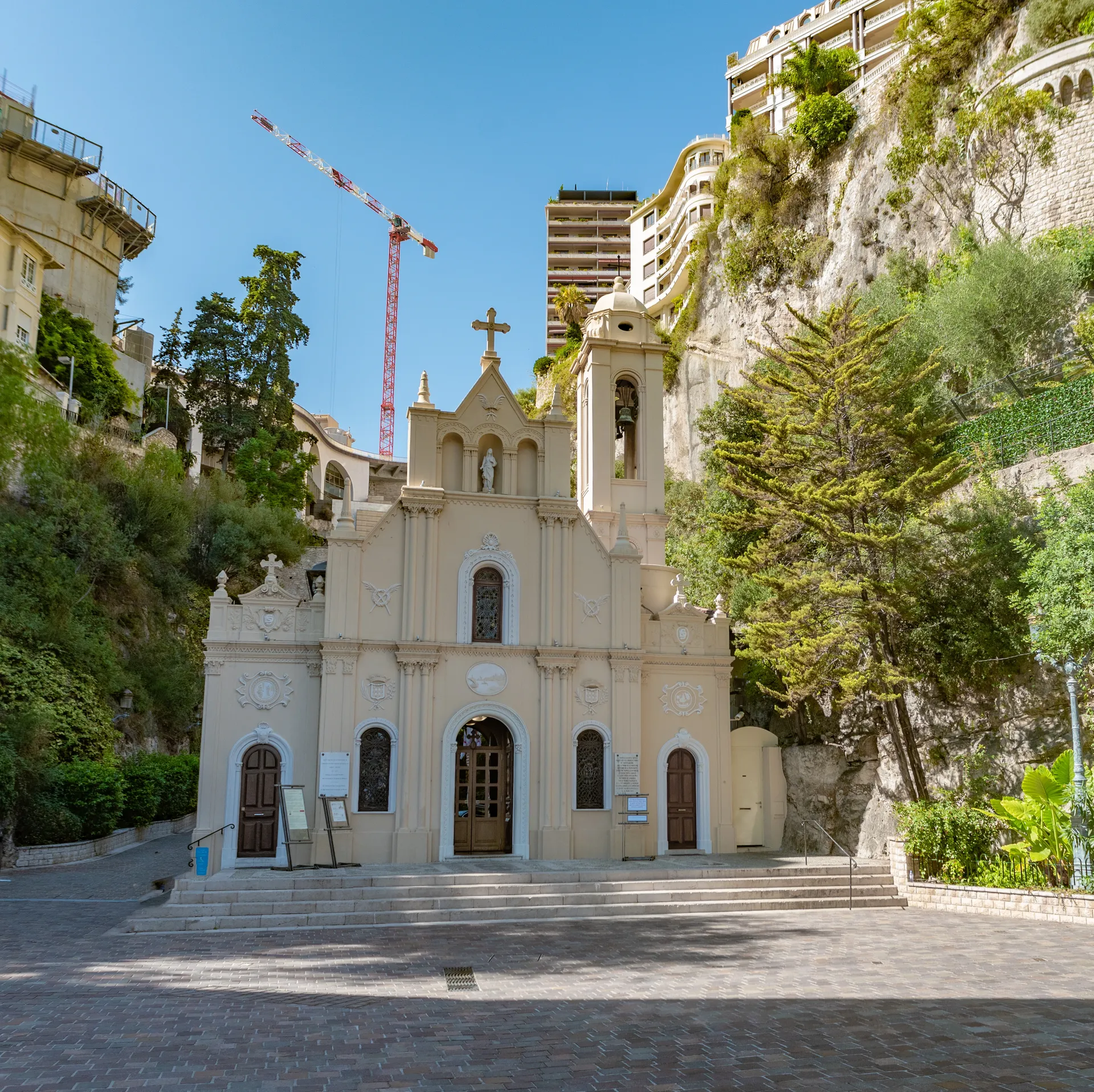 Small church in Monaco set against a rocky cliff with greenery and nearby urban development.