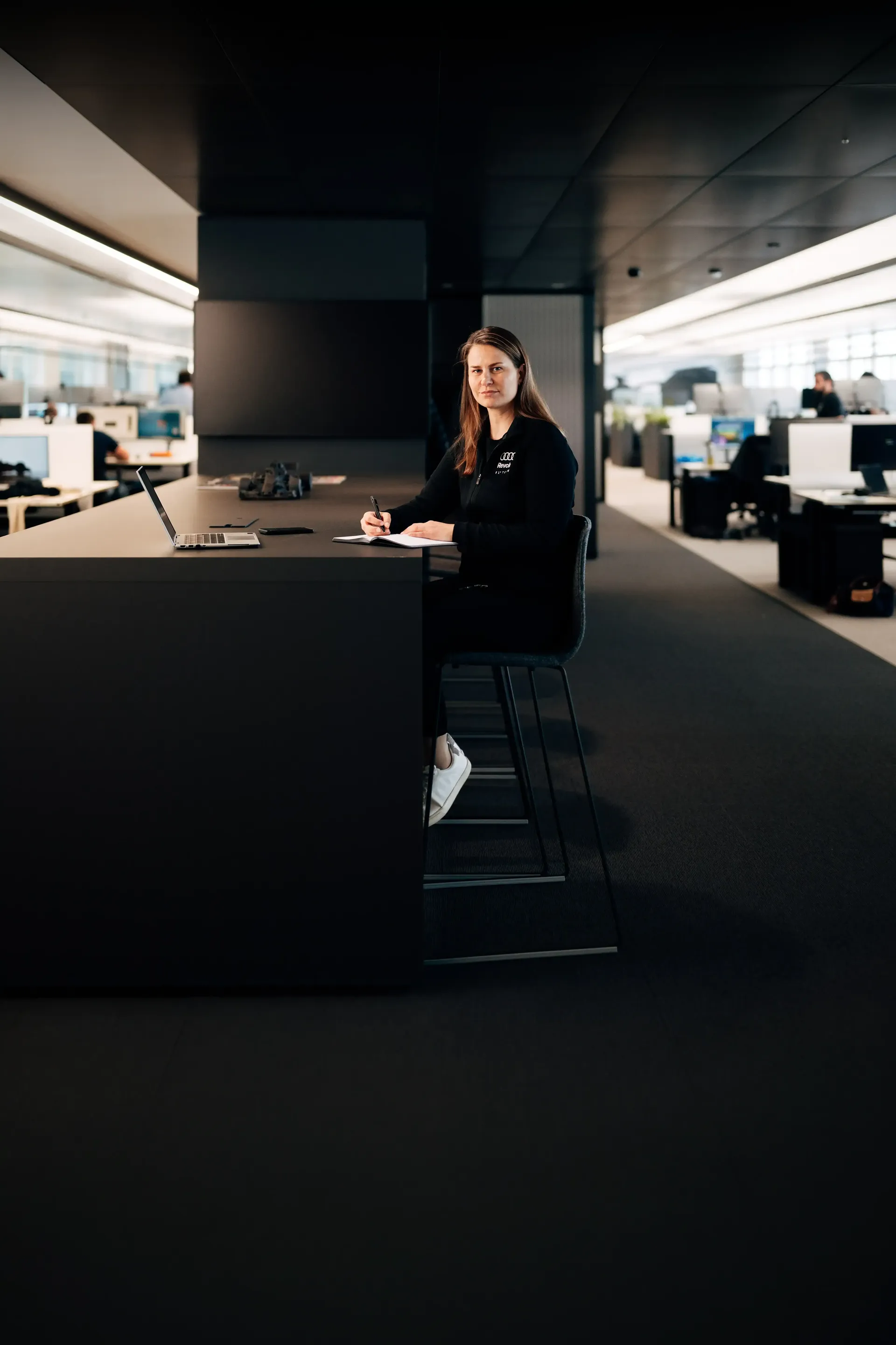 Sandra Internal communications of Audi Revolut F1® Team looking up from her desk, with a large office space behind her
