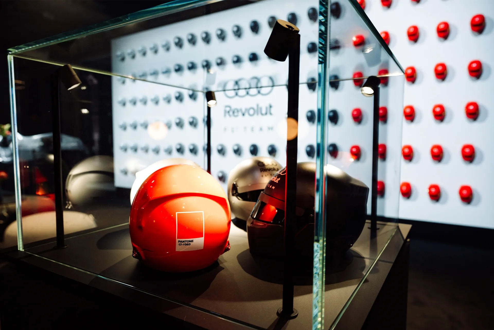 Glass display case featuring three modern helmets (red, silver, and blac  under spotlights, with a red Pantone-labeled helmet in the foreground and a patterned wall with red and dark circular elements in the background