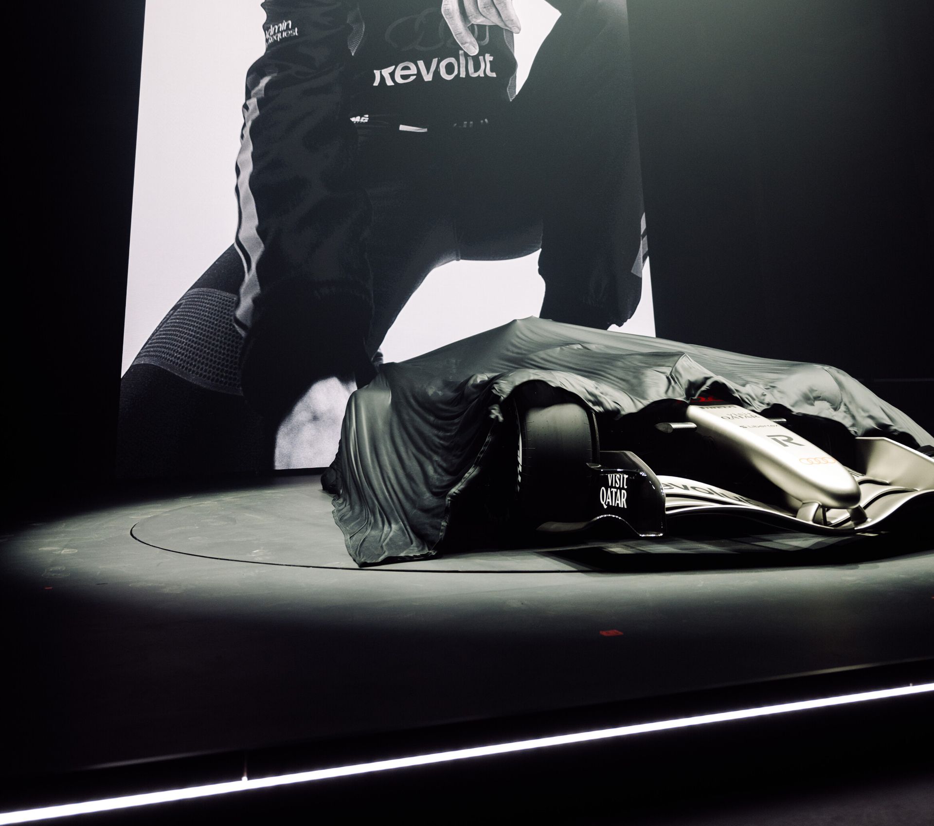 Formula 1 car partially covered by a draped cloth on a circular stage, with Visit Qatar branding visible and a large black-and-white screen of a driver with Revolut branding in the background