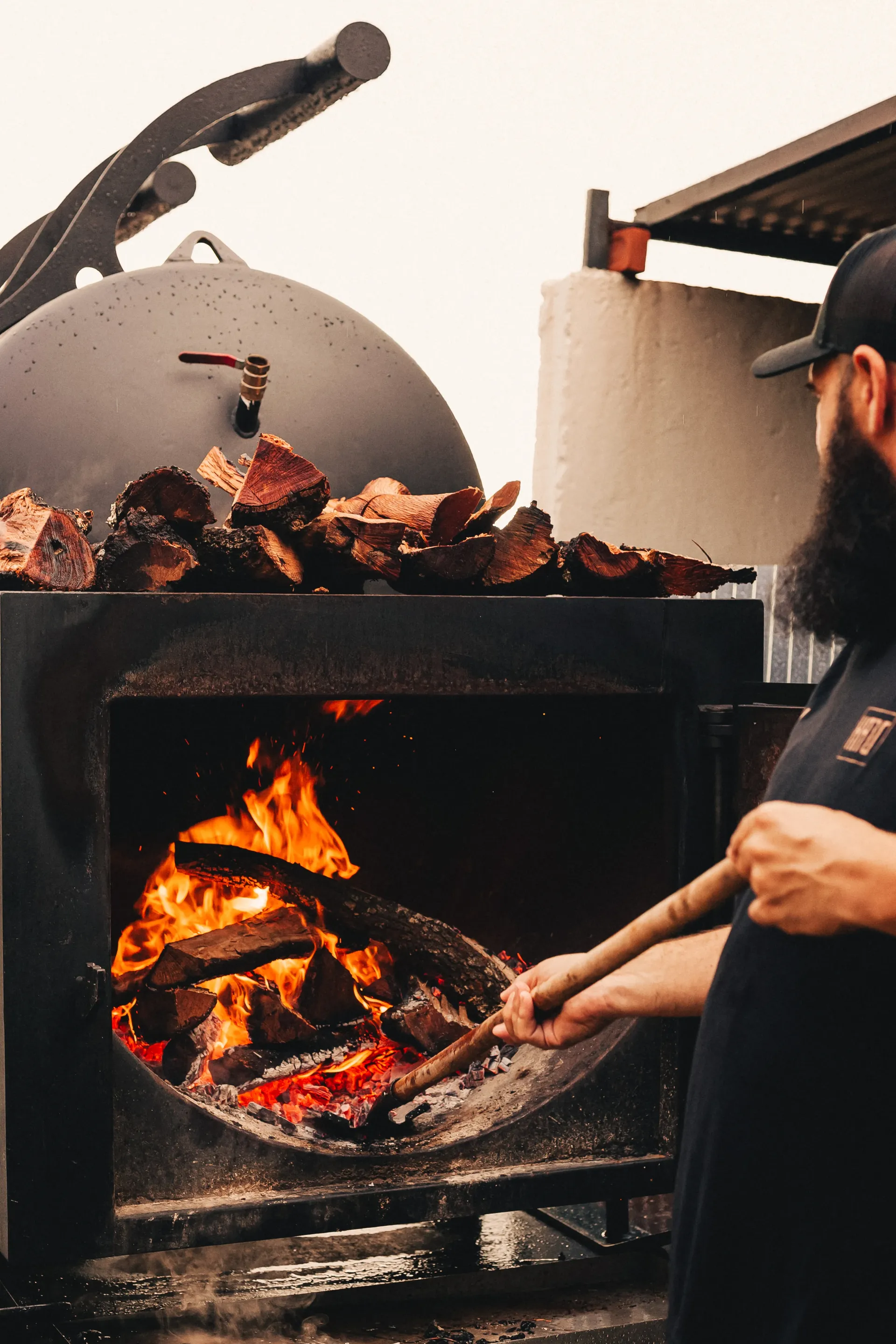 Pitmaster stoking the wood fire of a barbecue smoker with split logs stacked on top