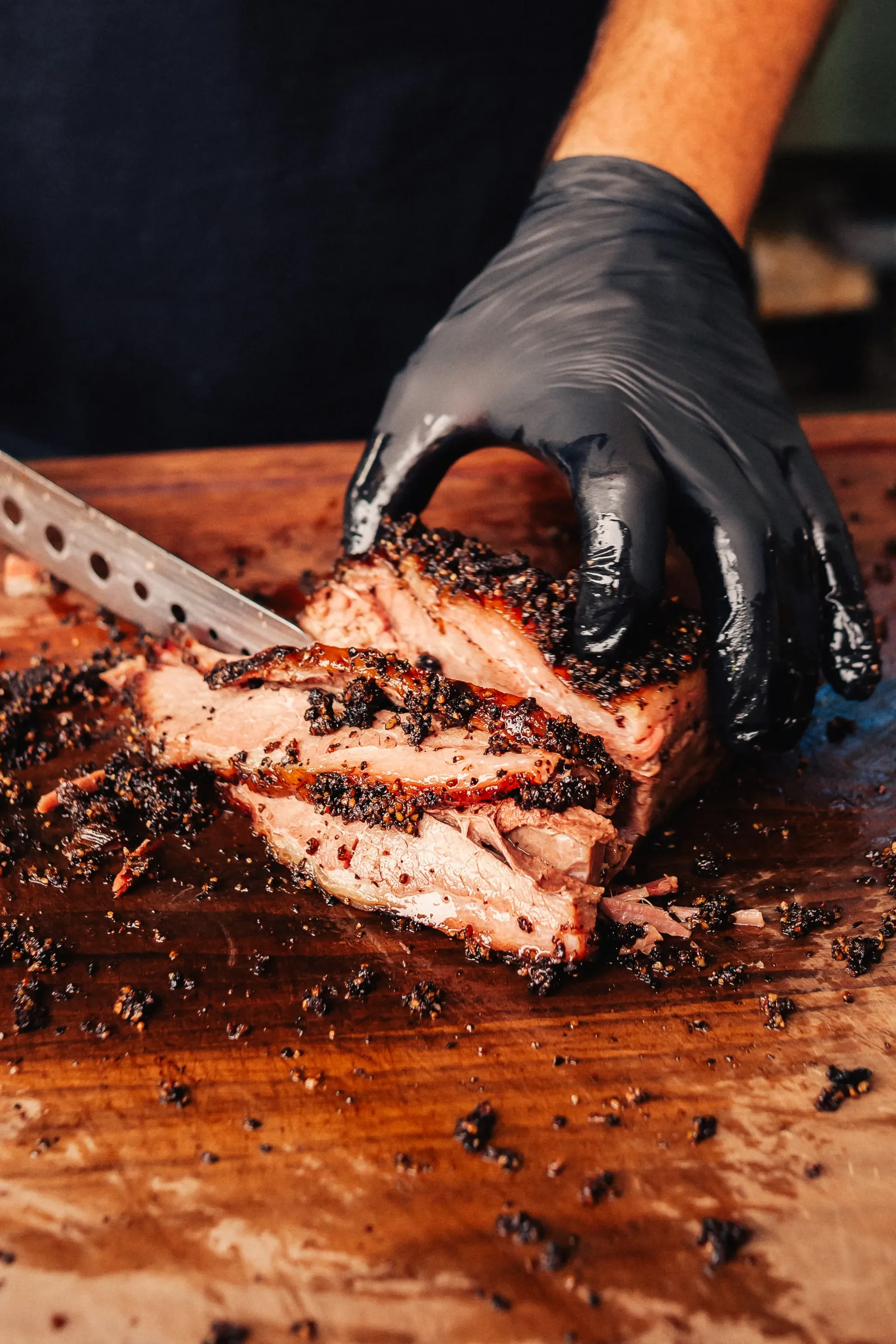 Gloved hand slicing smoked brisket with a peppery bark on a wooden cutting board