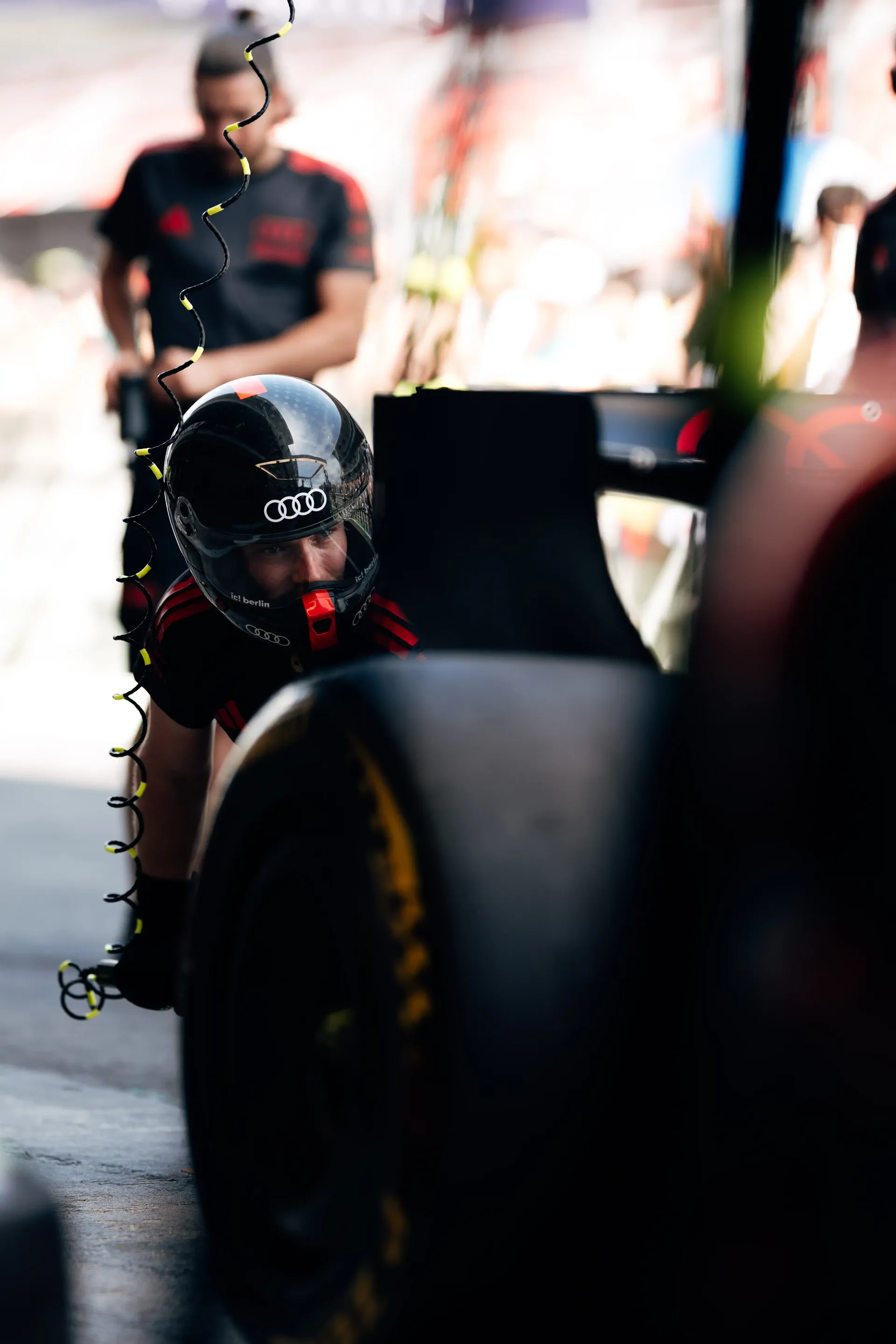 Pit crew member in helmet and gear leaning into the car during a stop, with blurred team activity in the background, Audi Revolut F1® Team.