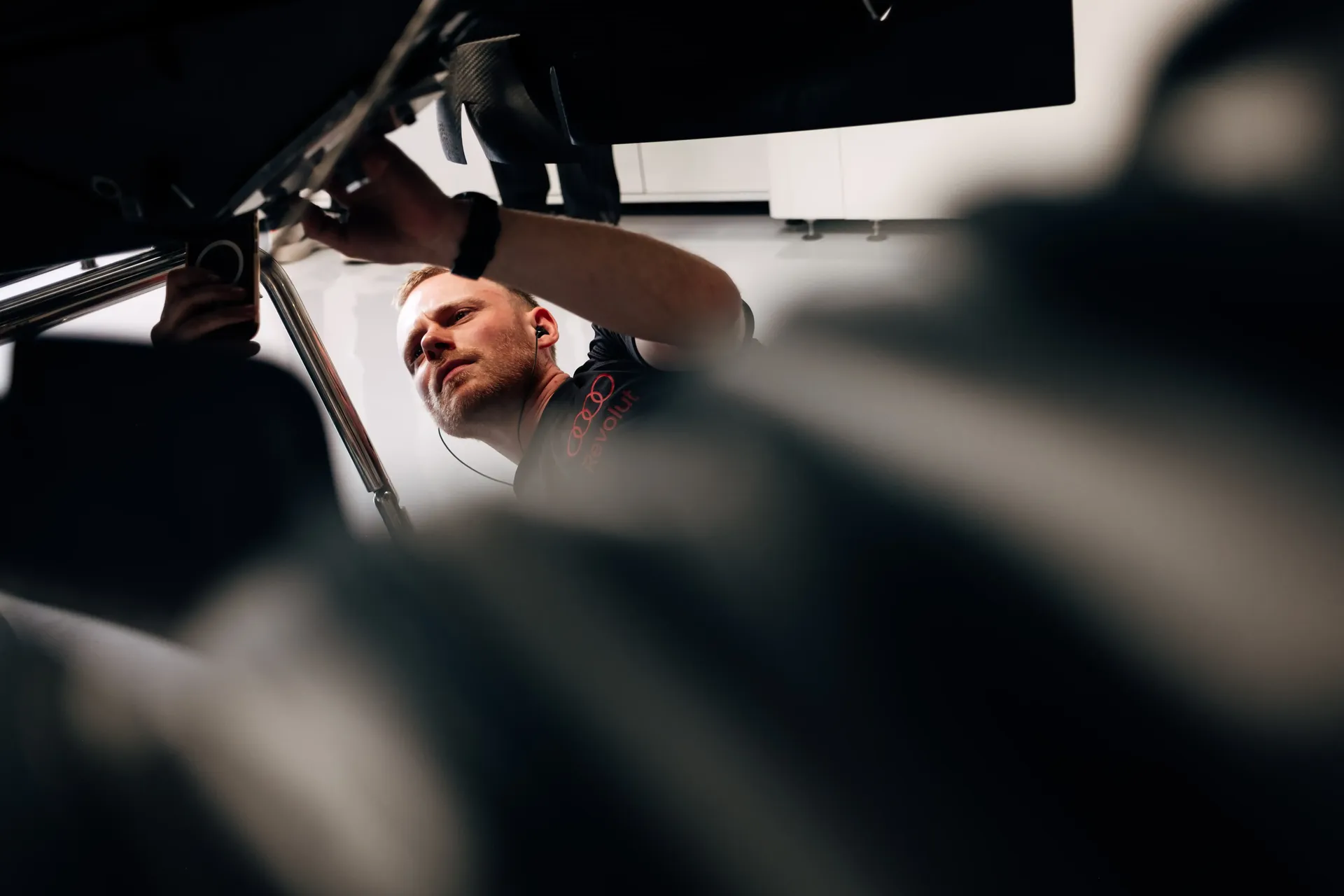 Dynamic angle from inside or beneath the car looking outward at a team member working overhead, conveying depth and mechanical intensity, Audi Revolut F1® Team.