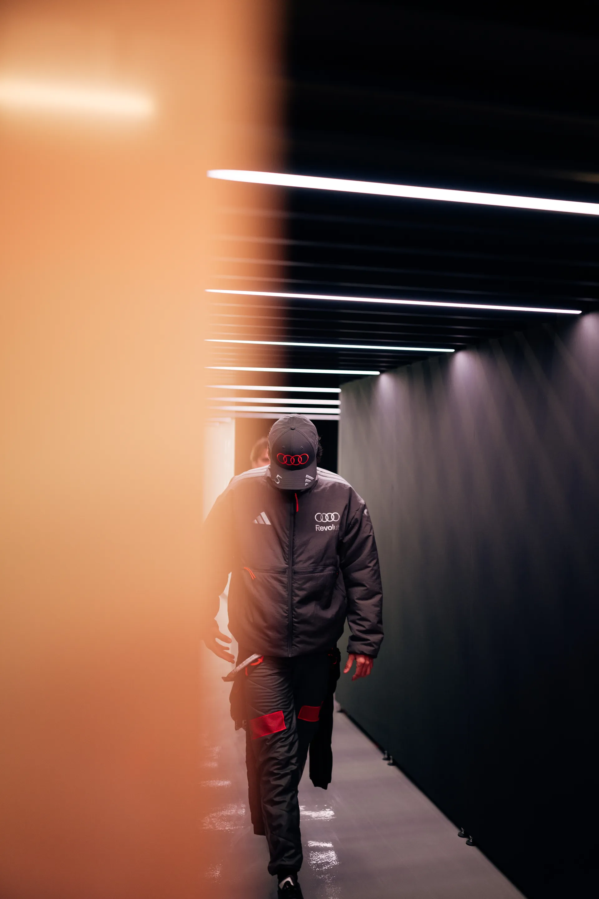 Gabriel Bortoleto walks through the Audi Revolut F1® Team garage tunnel in Shanghai, framed by warm foreground blur and overhead lights.