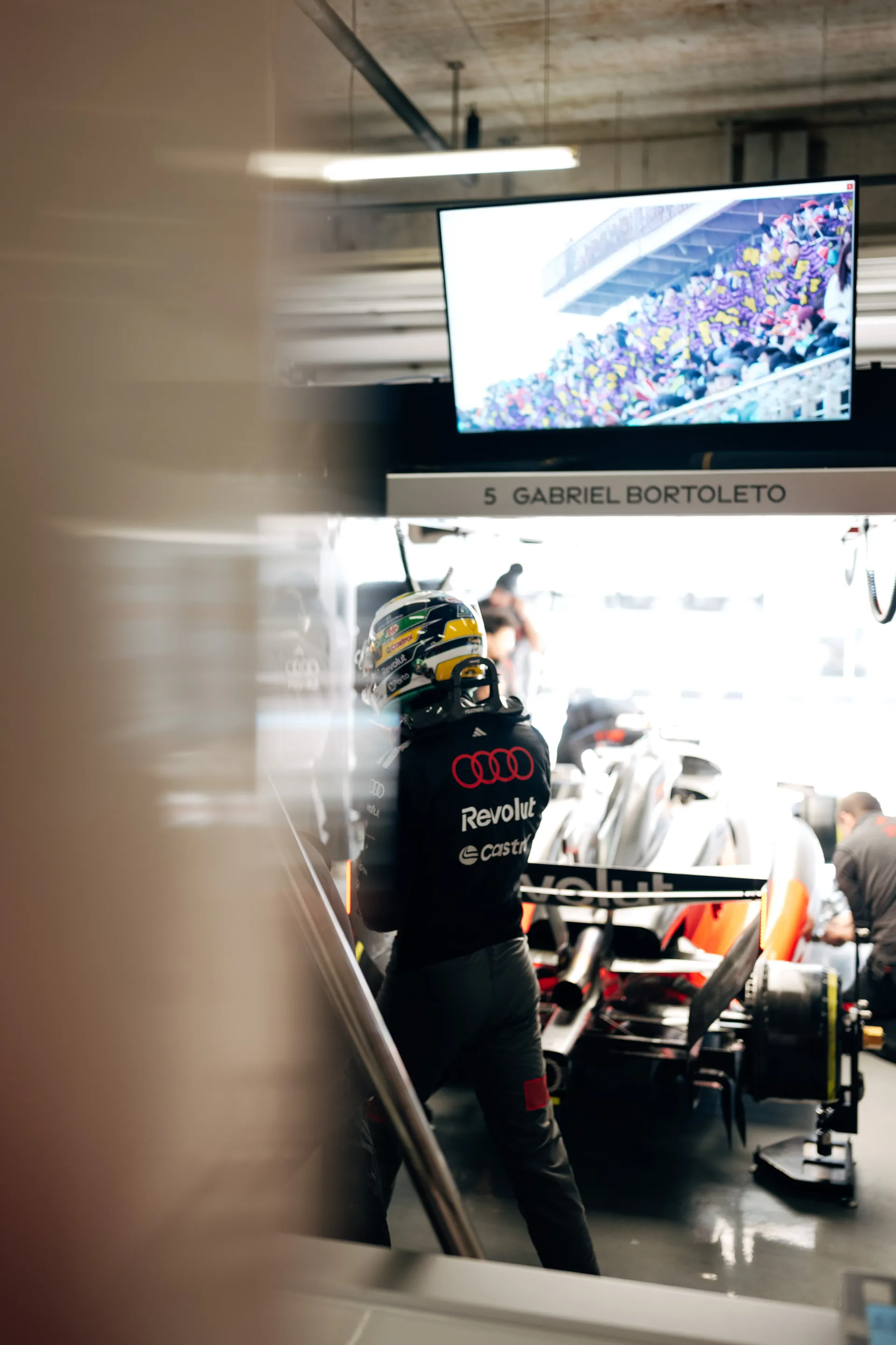 Gabriel Bortoleto stands beside his race car in the Audi Revolut F1® Team garage, with his pit bay screen visible above.