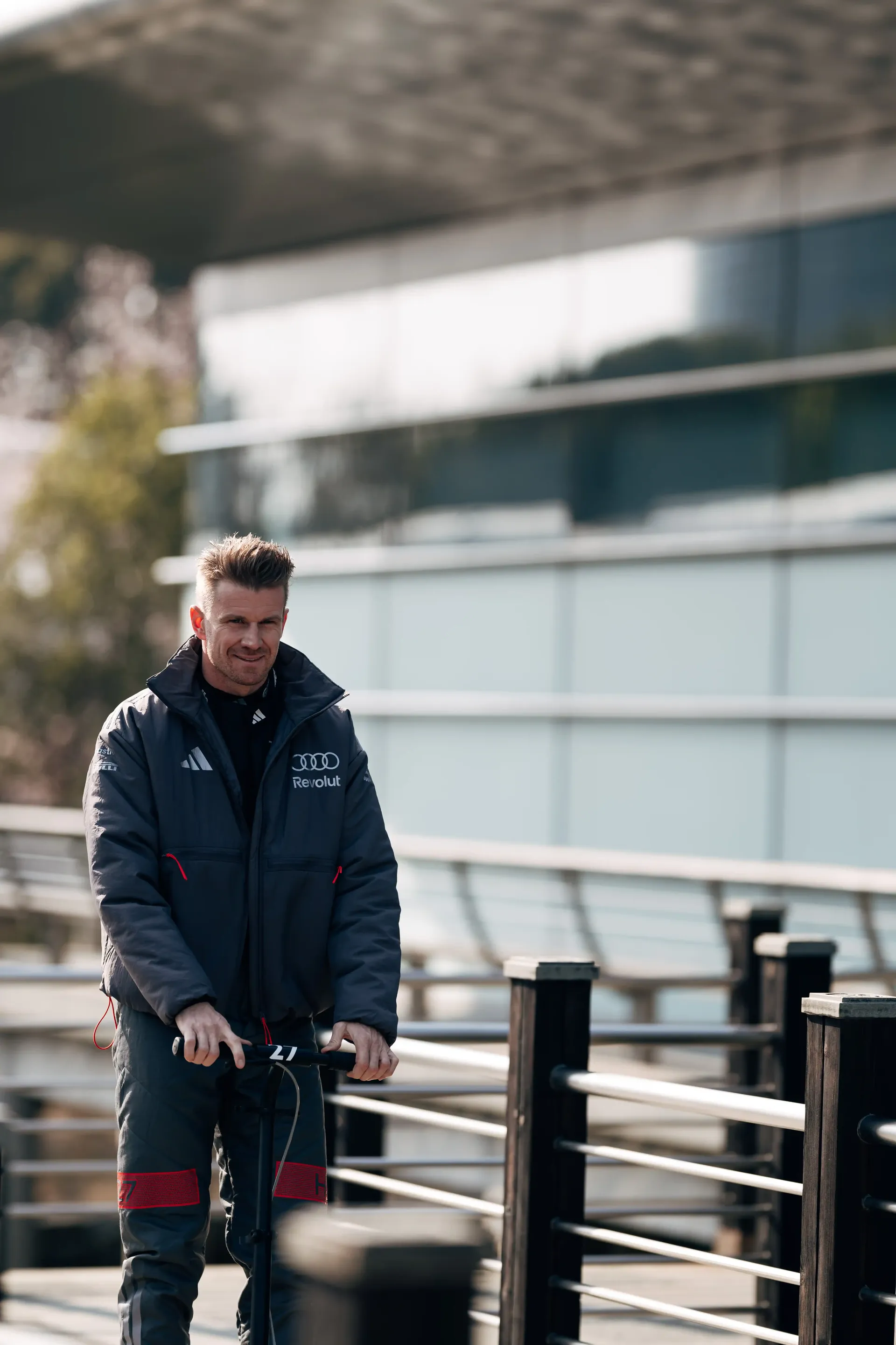 Audi Revolut F1® Team member Nico Hulkenberg riding an electric scooter along a paddock walkway near a modern racing facility.