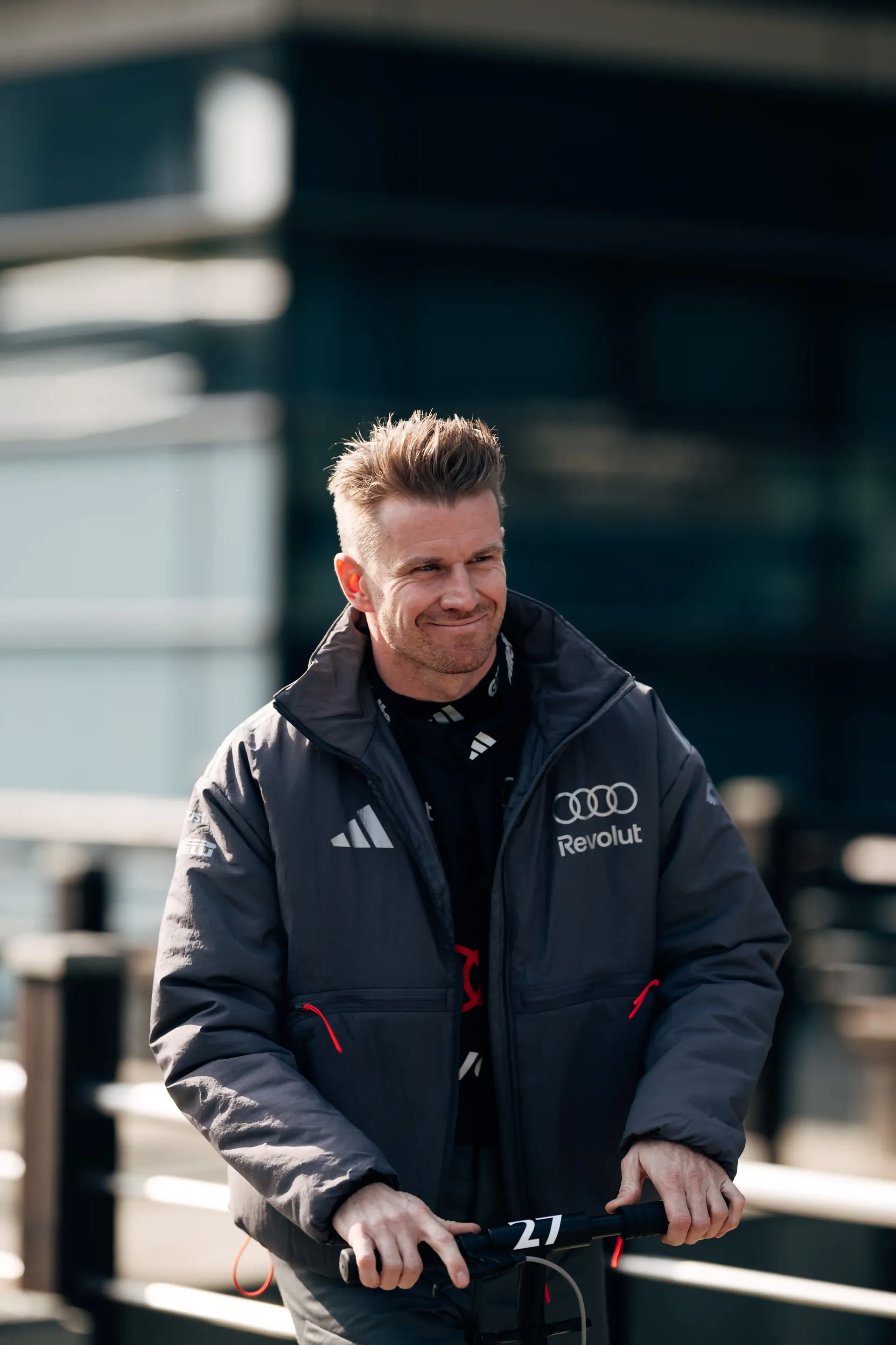 Smiling Audi Revolut F1® Team member Nico Hulkenberg riding an e-scooter outside the paddock area at a race track.