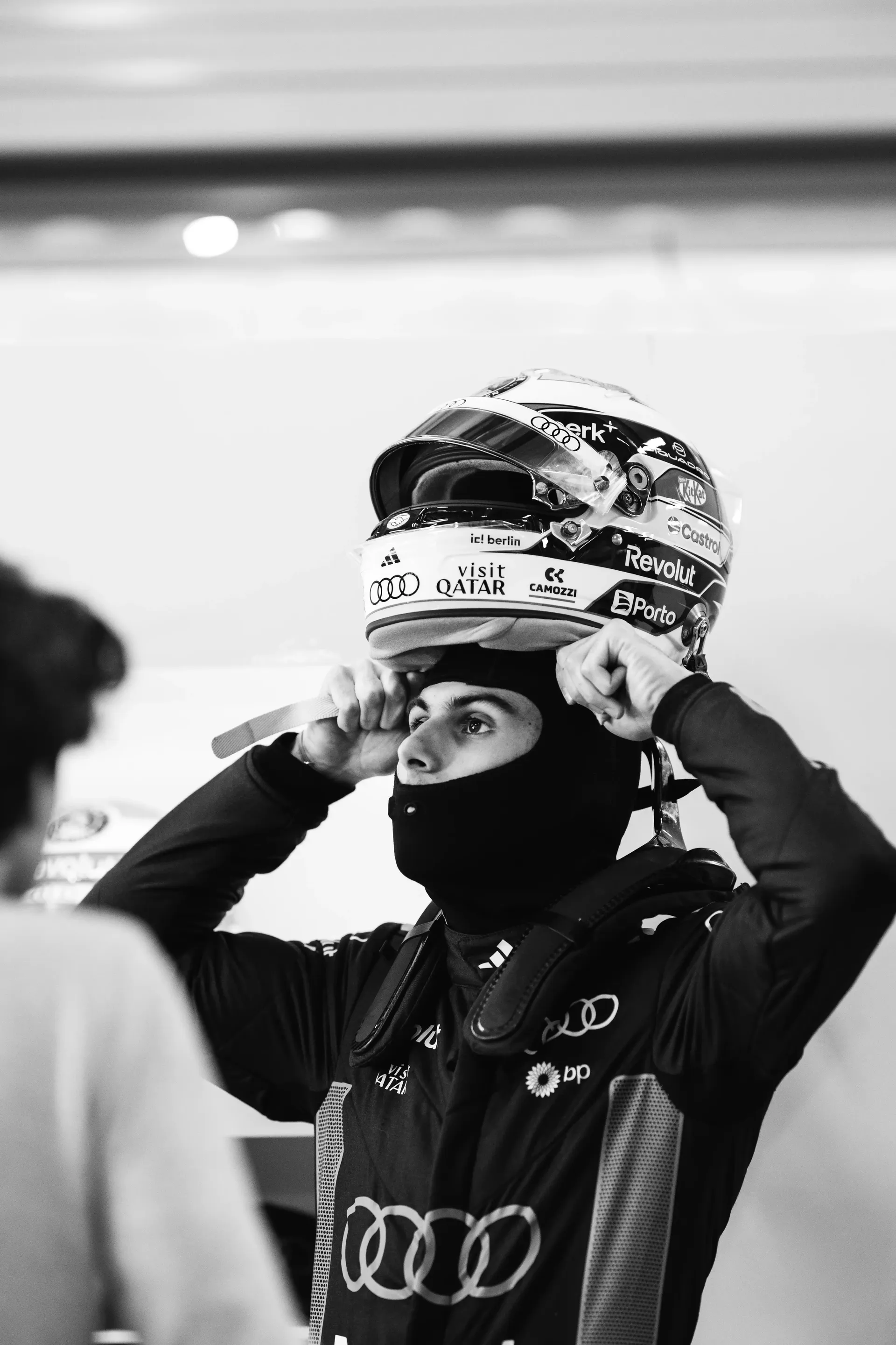 Black and white photo of an Audi Revolut F1® Team driver Gabriel Bortoleto adjusting his helmet and balaclava in the garage.