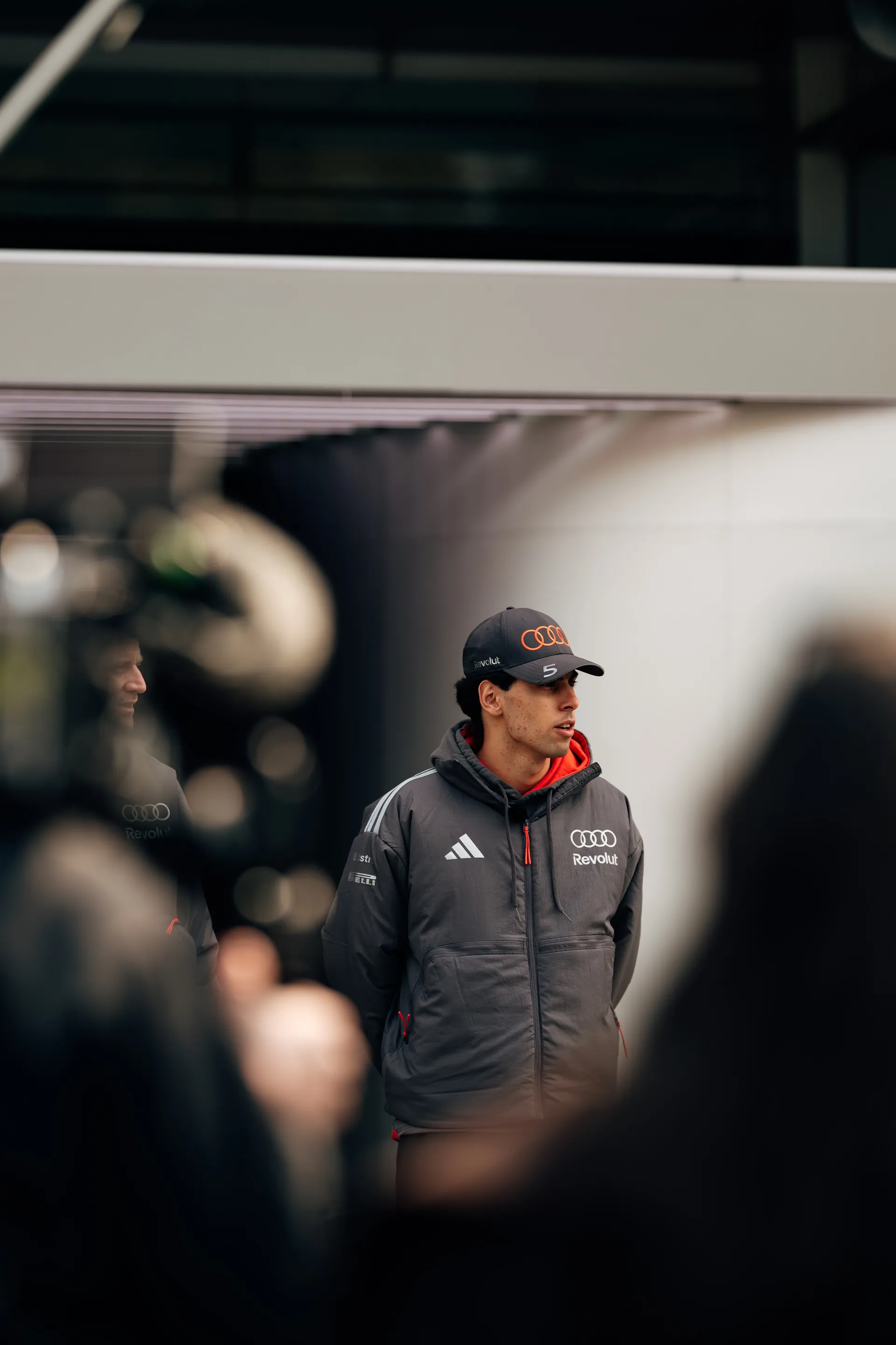 Gabriel Bortoleto stands outside the Audi Revolut F1® Team garage in Shanghai, framed by blurred foreground figures and equipment.