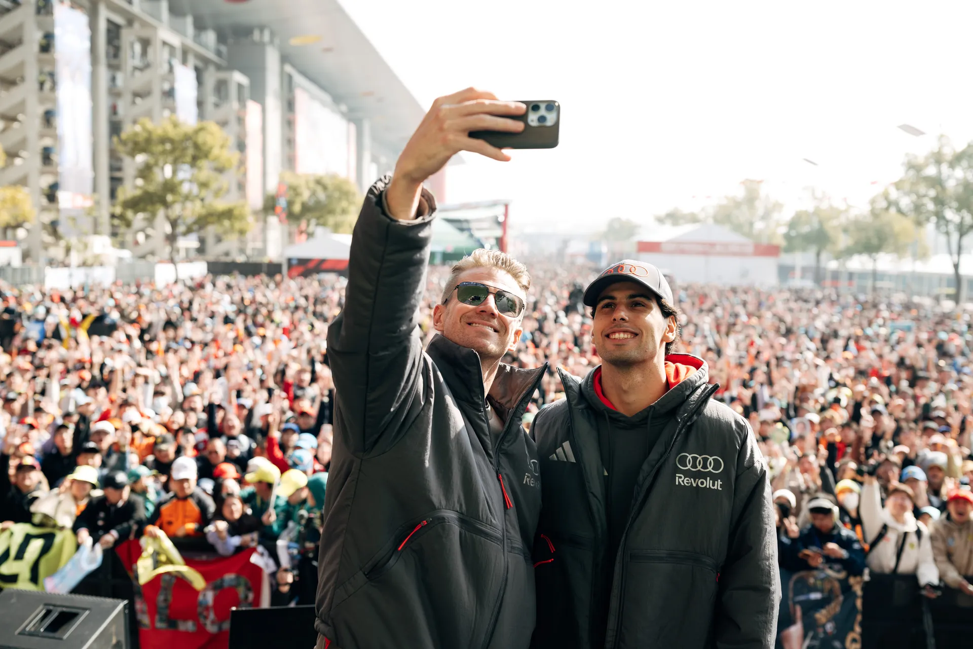 Nico Hulkenberg and Gabriel Bortoleto pose for a selfie on stage in front of a large Shanghai crowd at an Audi Revolut F1® Team appearance.
