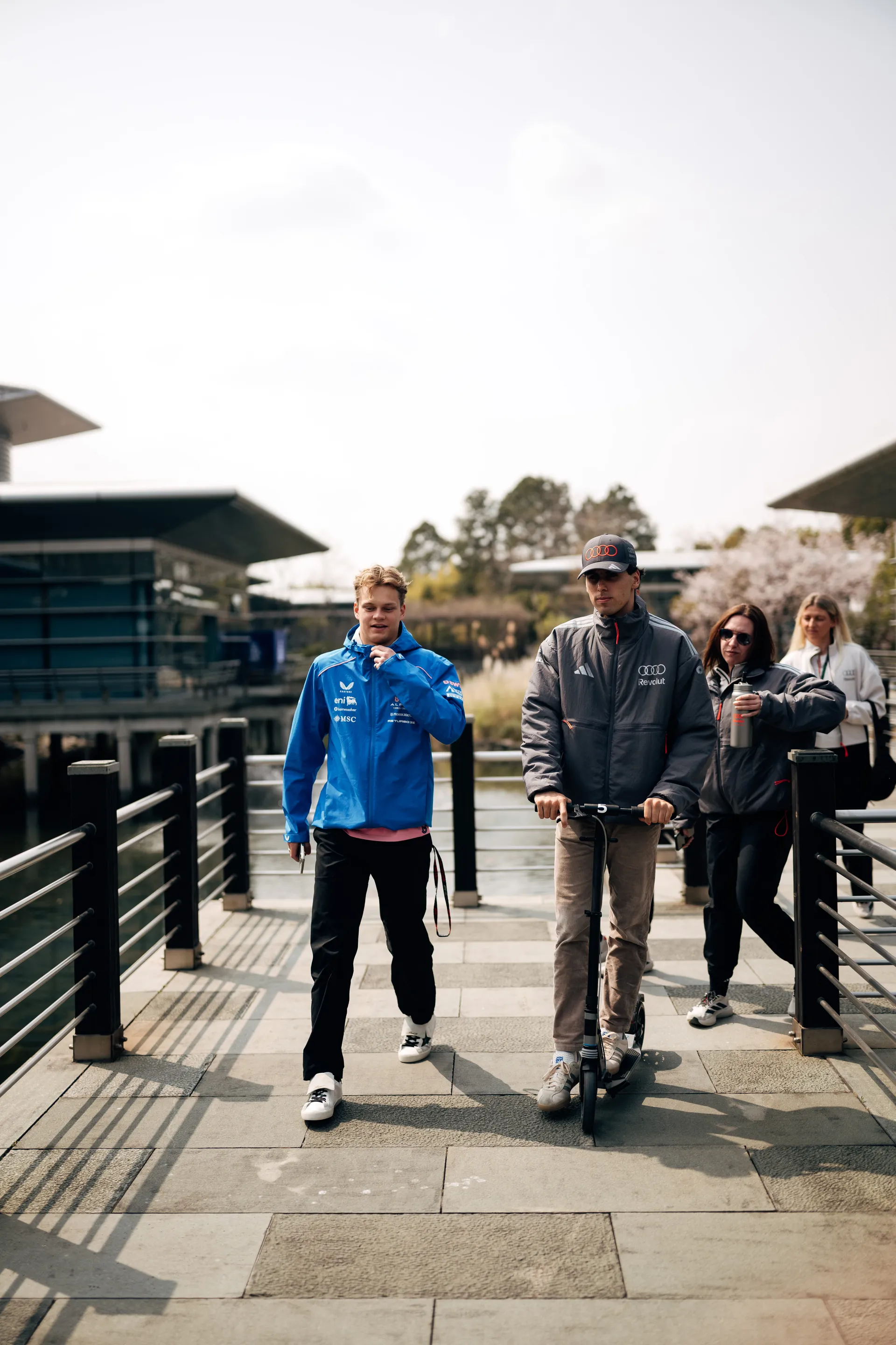 Gabriel Bortoleto rides across a bridge in the paddock on race day alongside team members and another driver.