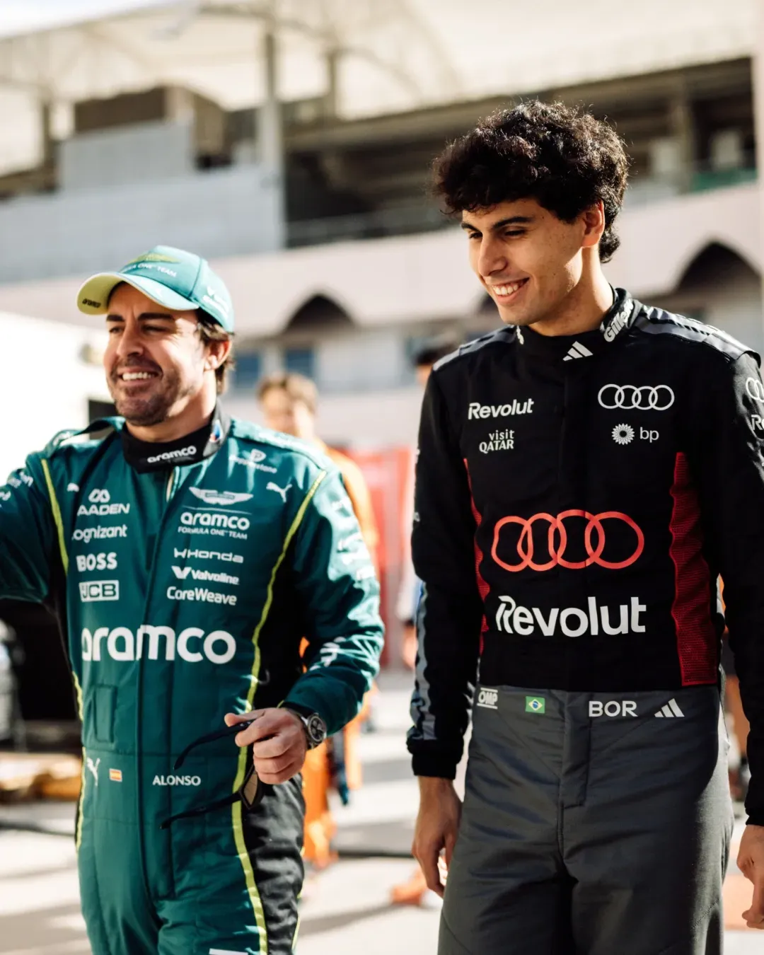 Two Formula 1 drivers Gabriel Bortoleto walk through the paddock, one in Audi Revolut F1® Team kit, smiling in Bahrain sunlight. Bahrain Testweek 2026 from Audi Revolut F1® Team