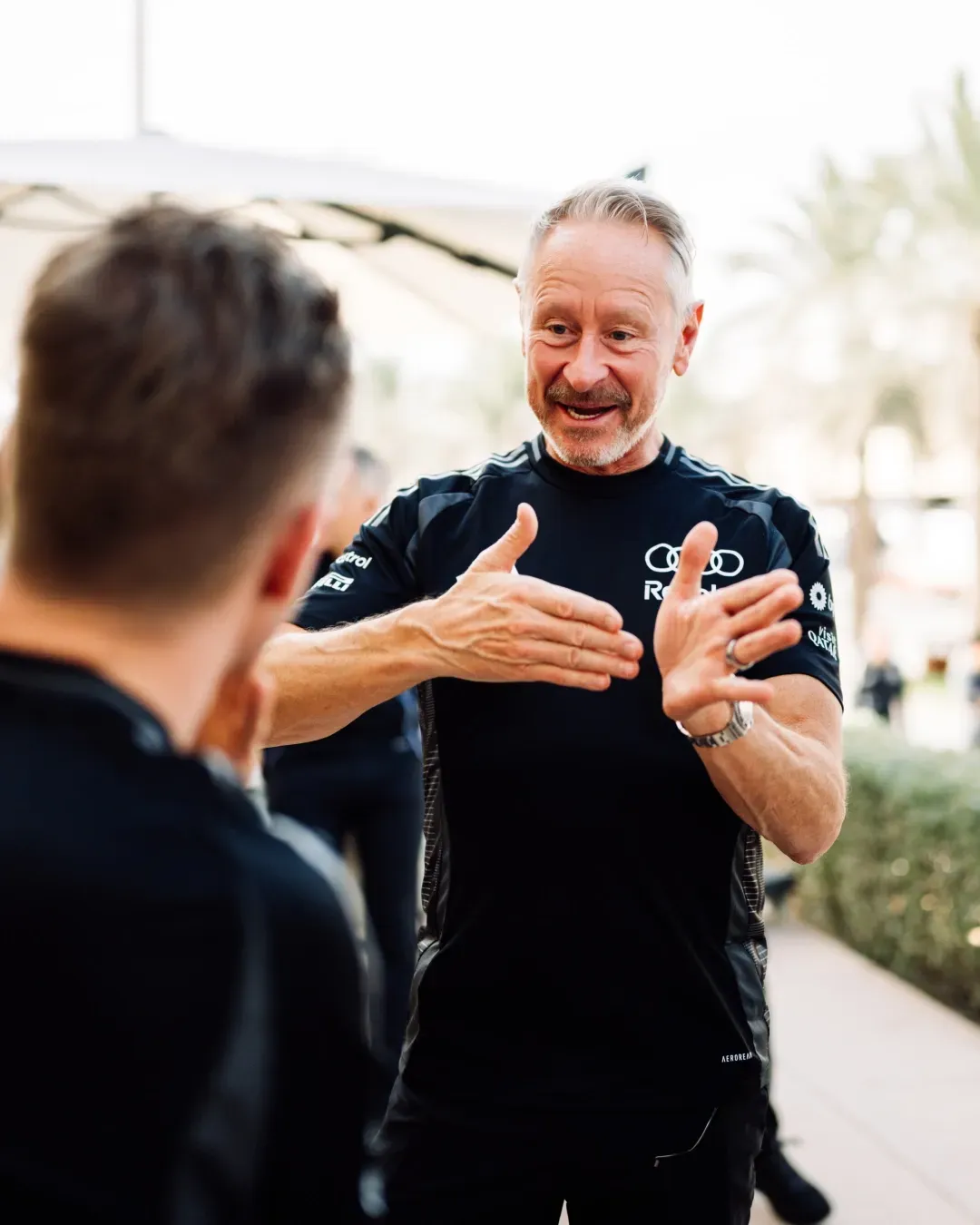 Audi Revolut F1® Team staff member briefs colleagues outdoors, hands mid gesture, palms and paddock in background.