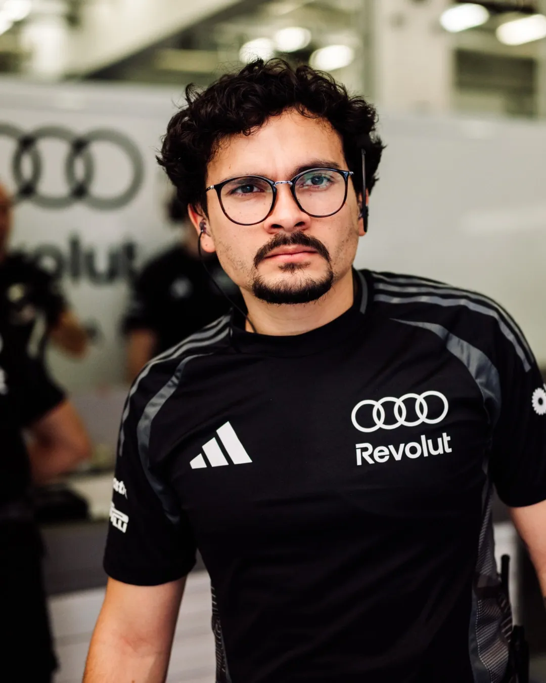 Portrait of an Audi Revolut F1® Team crew member in adidas shirt with glasses, focused expression inside the garage.