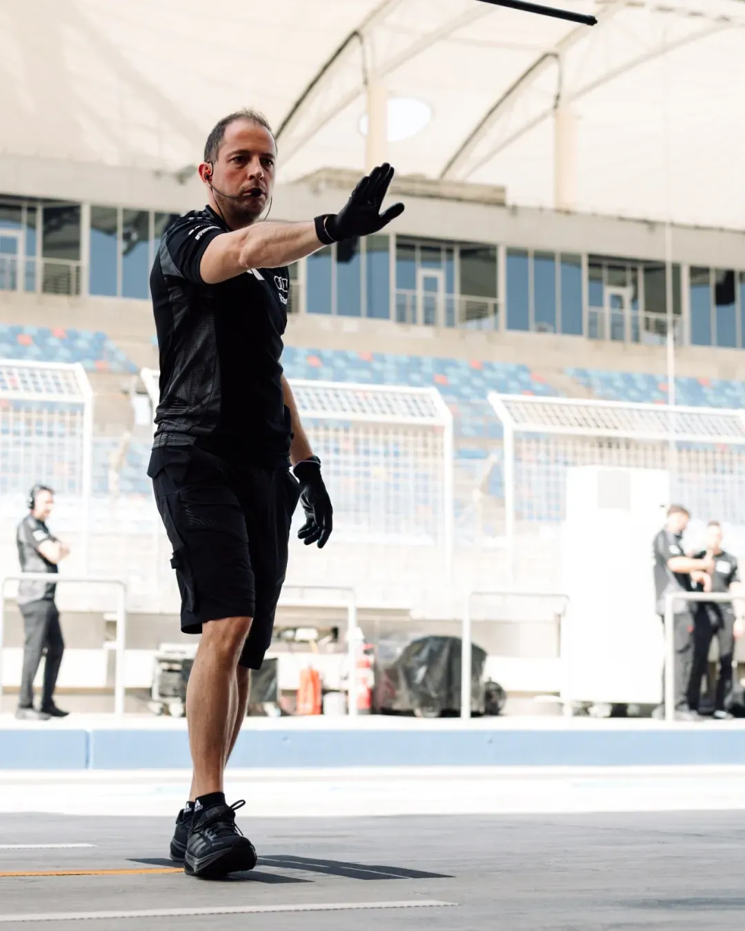 Pit crew member signals on the pit lane with a raised hand, grandstand and pit wall behind.