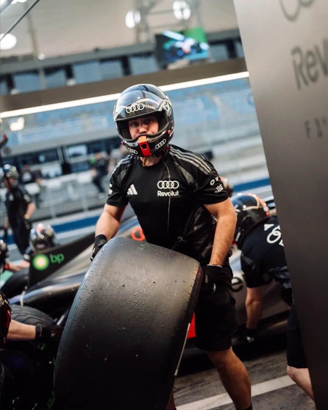 Pit crew member in helmet carries a slick tyre beside the car in the pit lane.