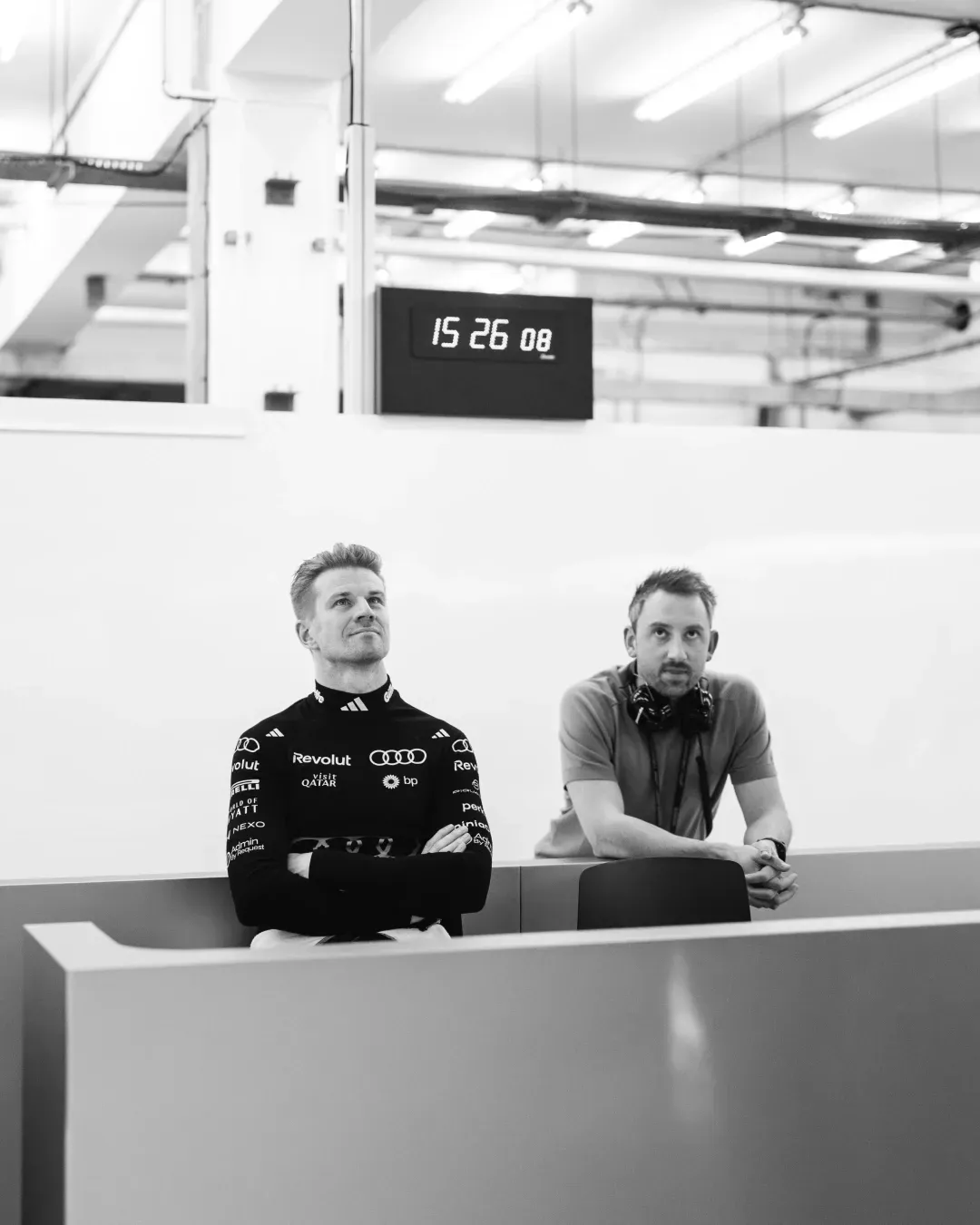 Two team members watch from the garage in black and white, digital clock above.