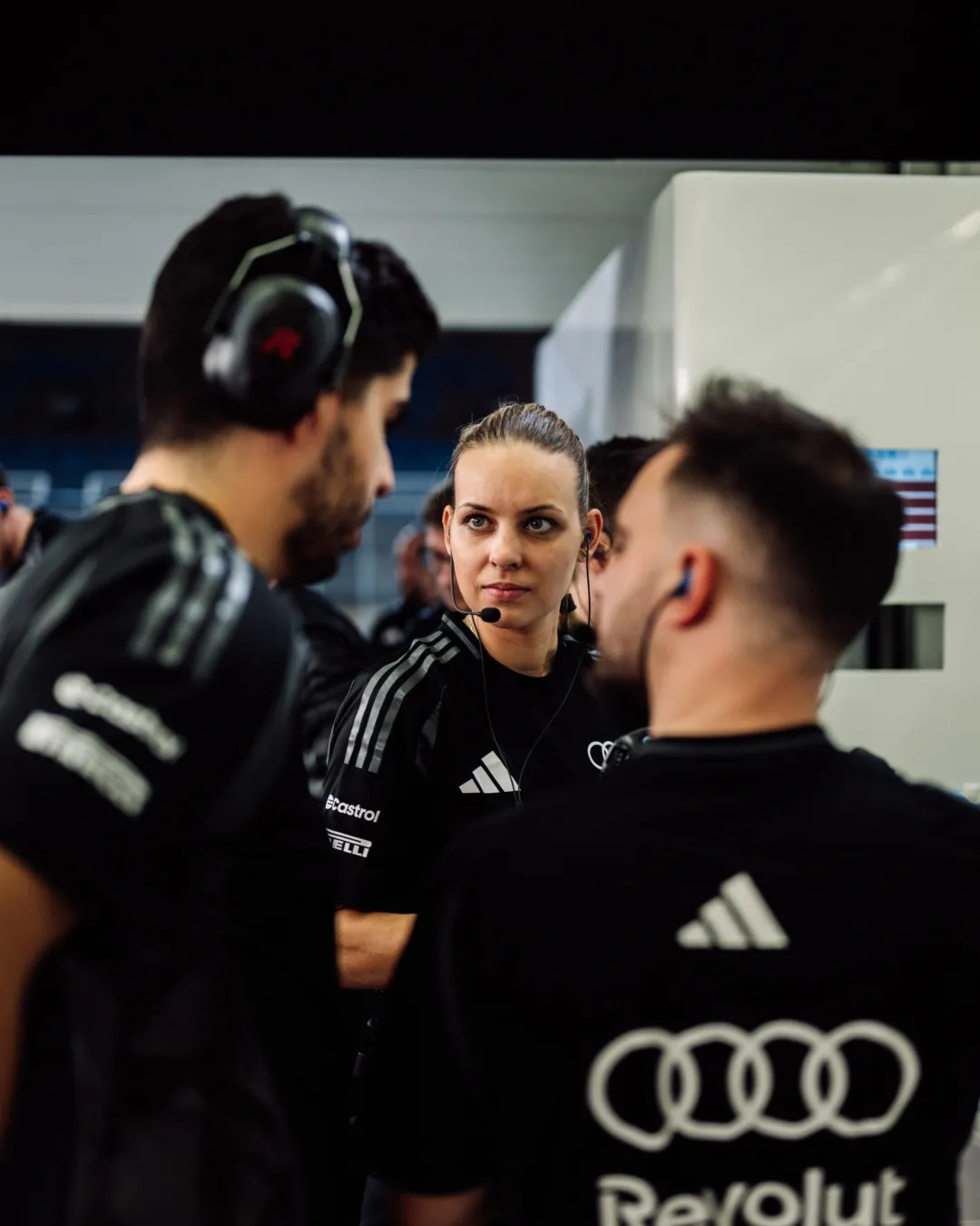 Race engineer with headset listens during a garage discussion, shown in black and white.