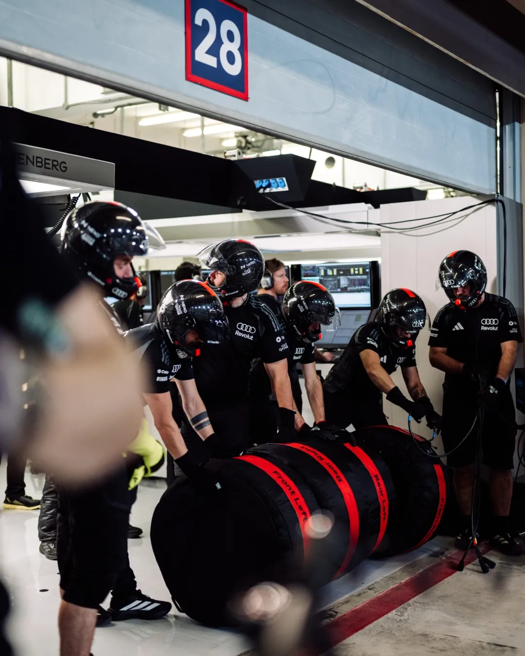Pit crew in helmets stand with stacks of tyres in garage bay 28.