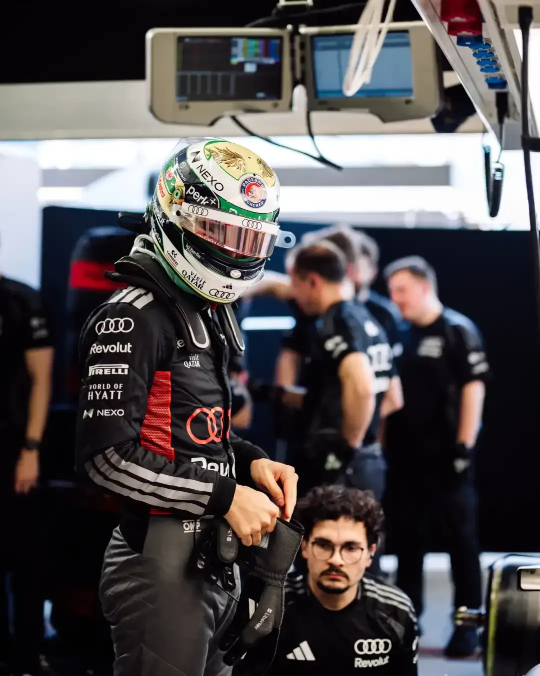Driver in full gear stands in the garage adjusting gloves while a mechanic crouches nearby.