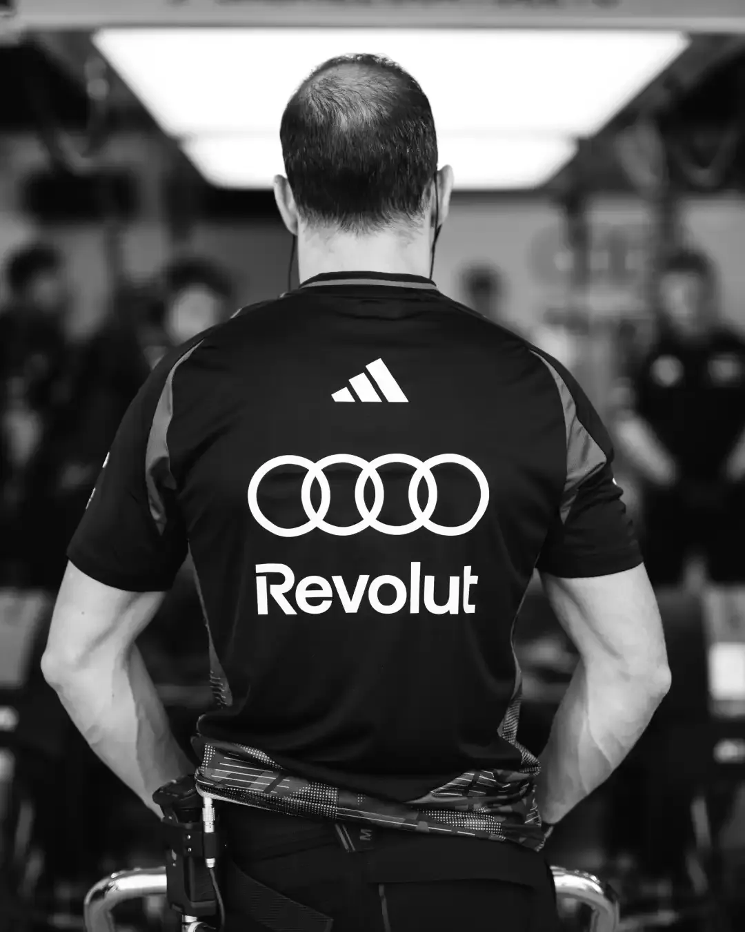 Black and white rear view of a team member wearing an Audi and Revolut branded shirt inside the garage.