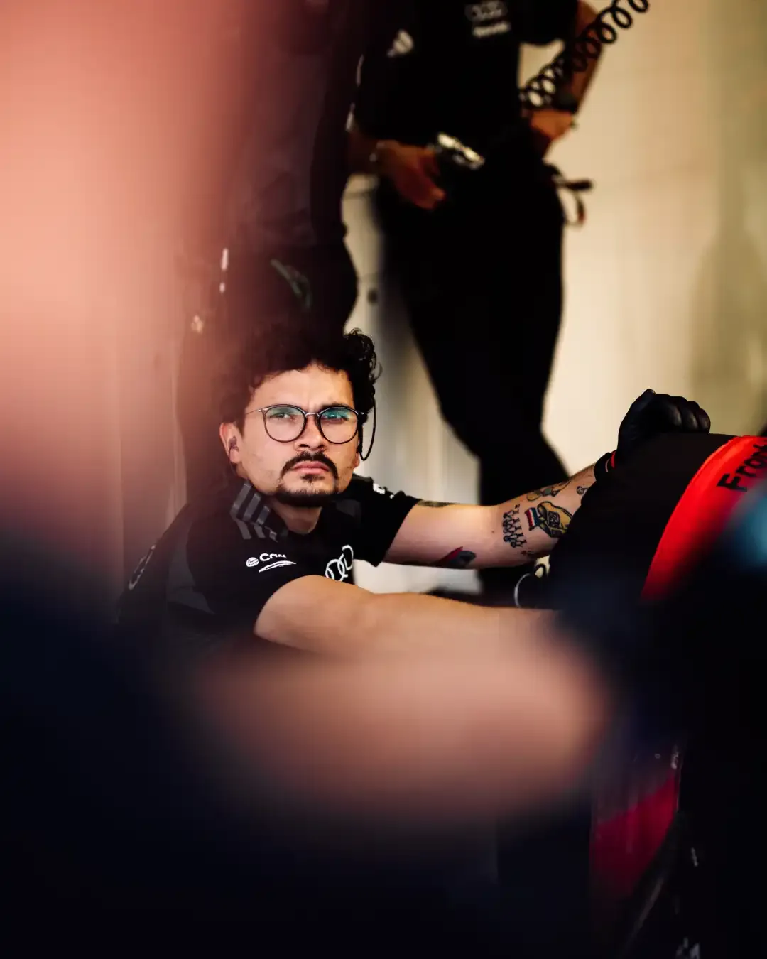 Mechanic with glasses sits beside a tire in the garage, watching the work area.