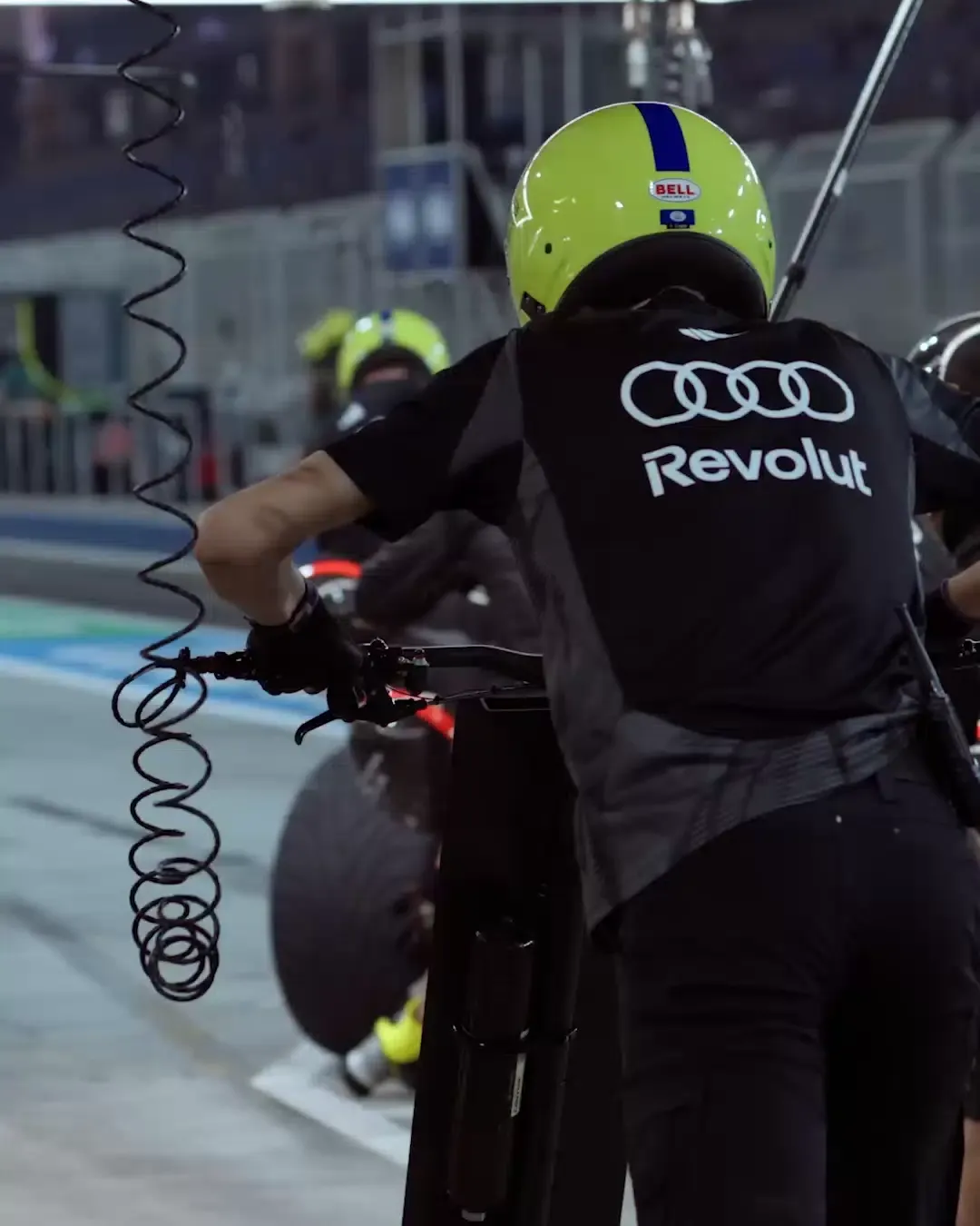 Audi Revolut F1® crew member waits for car to enter the Stop Pit, yellow neon Helmet, Closeup black Tshirt with Revolut and Audi Logo, car drives into the pitstop, ready for tire change