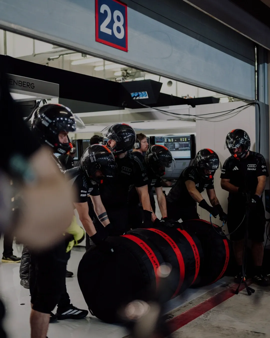 Pit crew in helmets stand with stacks of tyres in garage bay 28.