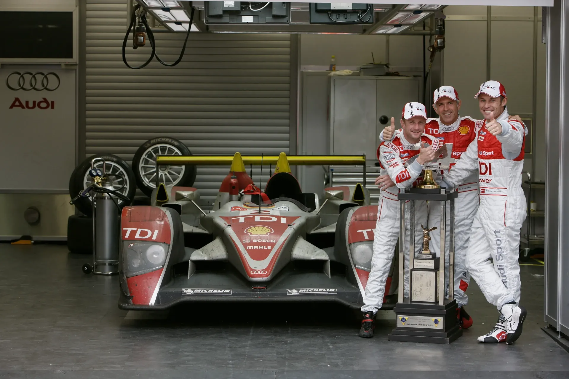 Tom Kristensen and Allan McNish stand beside an Audi Sport LMP1 race car in the garage with teammates.